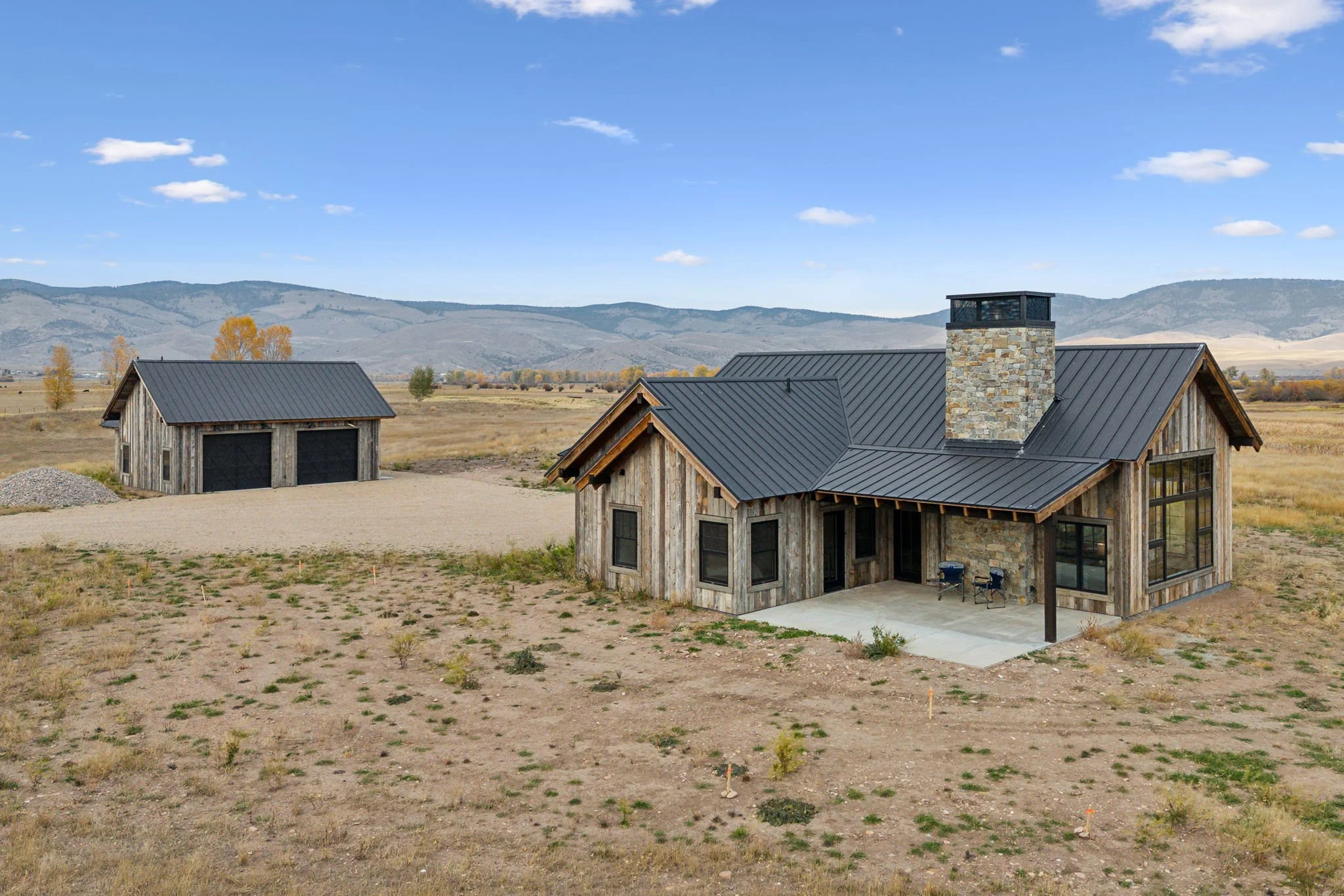 A modern farmhouse with a metal roof and stone chimney, located in a rural, open landscape with distant mountains in the background. Custom Home built in Montana by Edgell Building.