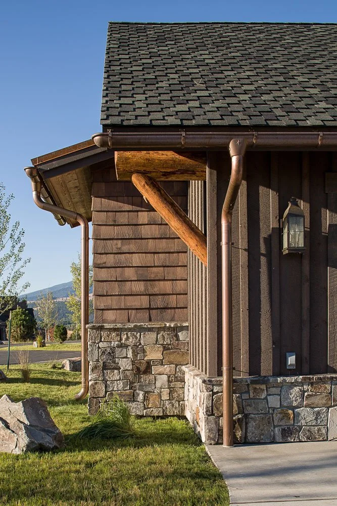 Close-up of a house corner with a stone foundation, wooden siding, and a shingled roof, featuring copper gutters and a wall-mounted outdoor lantern. Custom Home built in Montana by Edgell Building.