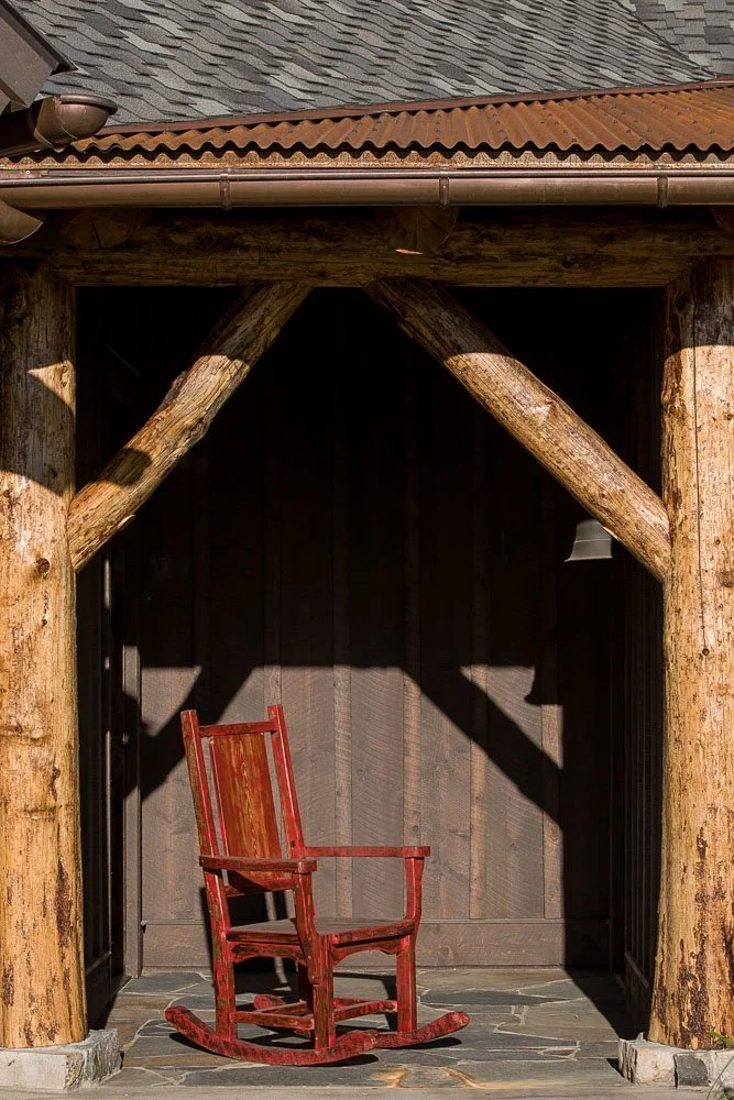 A red wooden rocking chair on a stone floor against a dark wooden wall, framed by large rustic wooden beams, with a textured roof above. Custom Home built in Montana by Edgell Building.