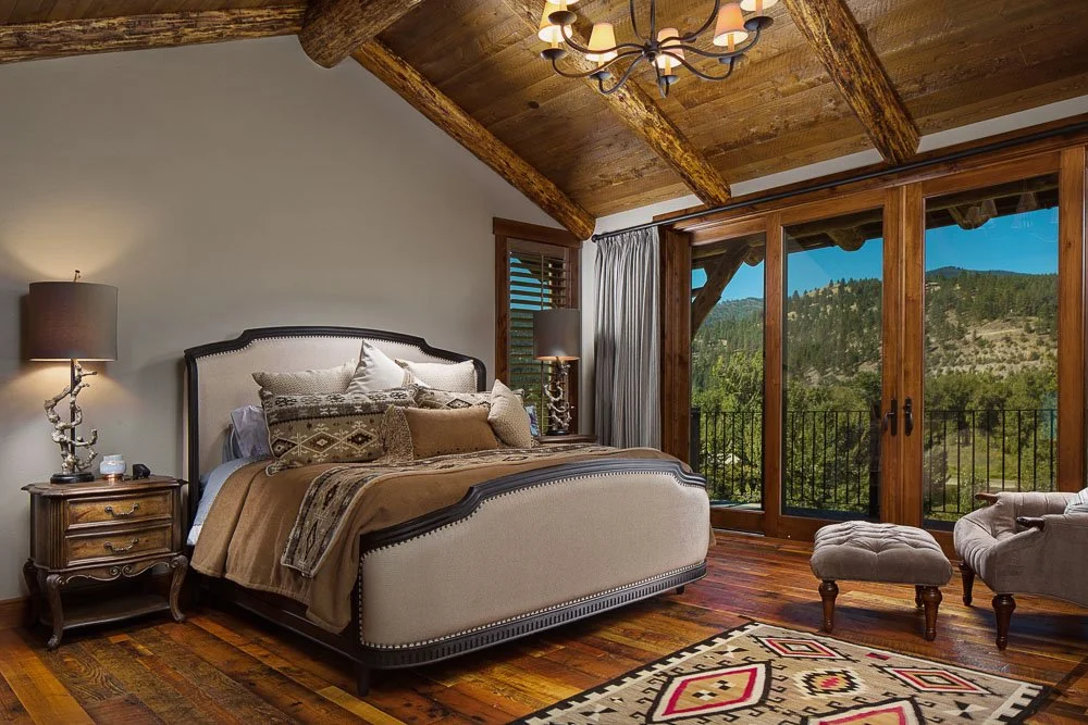 A bedroom with a large bed, wooden bedside tables with lamps, a cushioned armchair and ottoman, a large glass door overlooking a mountain landscape, wooden ceiling beams, and a patterned area rug. Custom Home built in Montana by Edgell Building.
