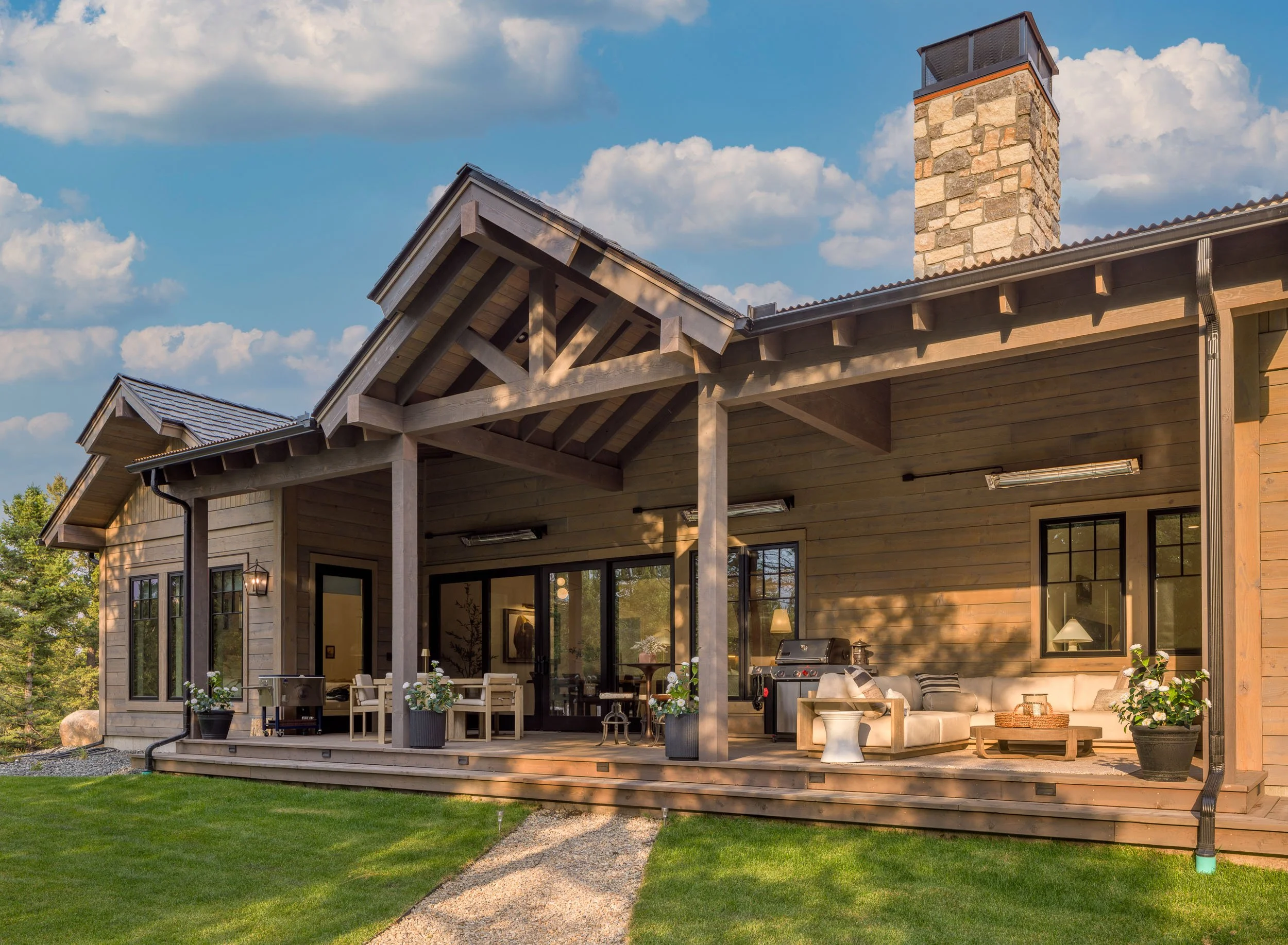A wooden house with a spacious outdoor patio, potted plants, outdoor seating, a barbecue grill, and a stone chimney, set against a backdrop of a blue sky with clouds and green lawn.