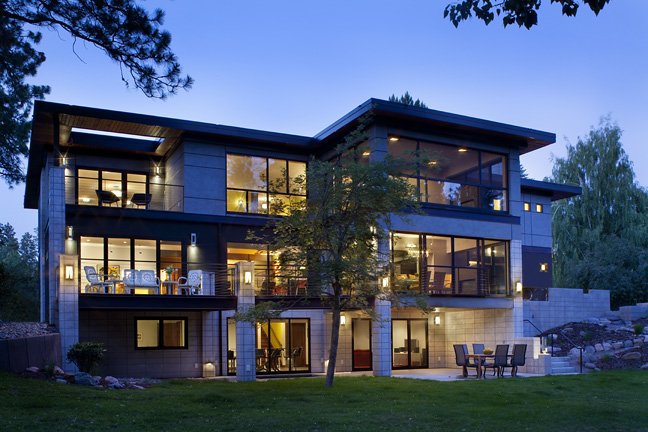 Modern multi-story house with large windows, outdoor patio, and landscaped yard during evening. Custom Home built in Montana by Edgell Building.