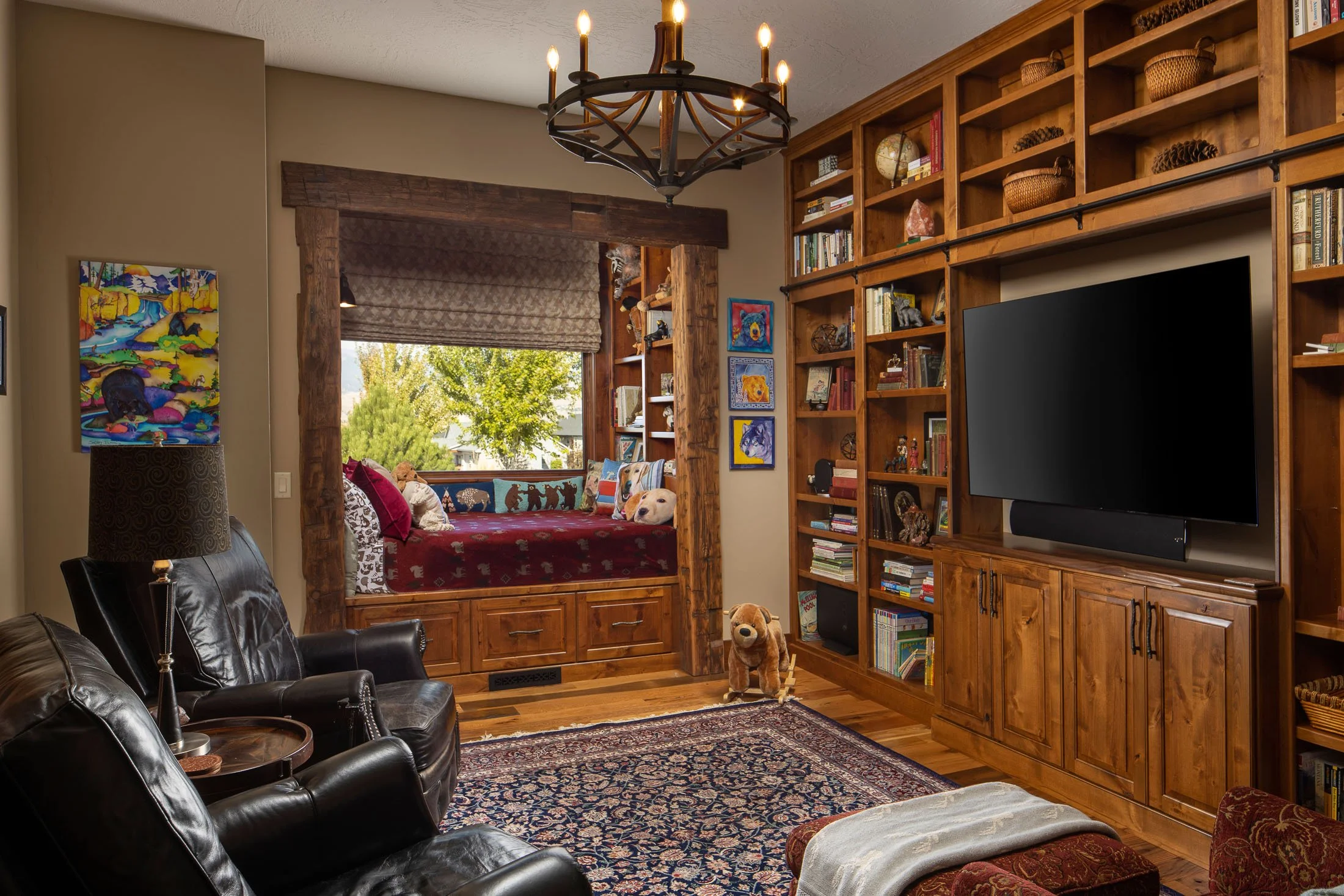Living room with black leather chairs, a large wooden bookshelf, a wall-mounted flat screen TV, a colorful area rug, and a window seat with pillows and a view of trees outside. Custom Home built in Montana by Edgell Building.