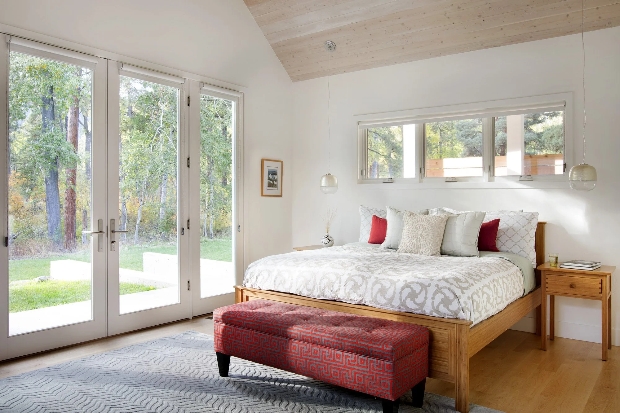 Bright bedroom with large glass double doors leading to an outdoor yard, a wooden bed with white patterned bedding, and a red upholstered bench at the foot of the bed. Custom Home built in Montana by Edgell Building.