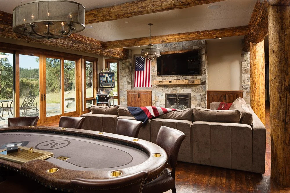 Living room with large sectional sofa, stone fireplace, flat-screen TV, an American flag, and a poker table with chairs, overlooking a deck with outdoor seating and a rural landscape. Custom Home built in Montana by Edgell Building.