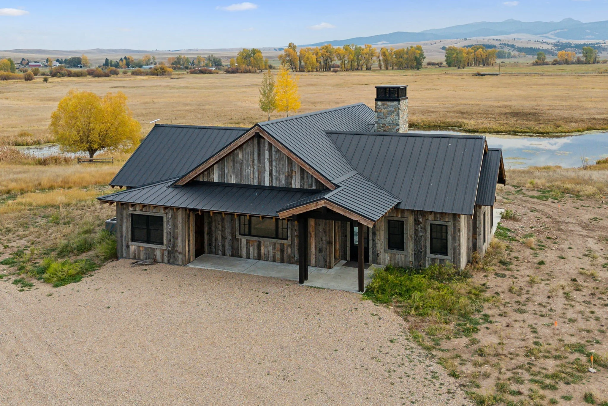 A rustic house with a metal roof in a rural landscape with grass, trees, and a pond, mountains in the background. Custom Home built in Montana by Edgell Building.