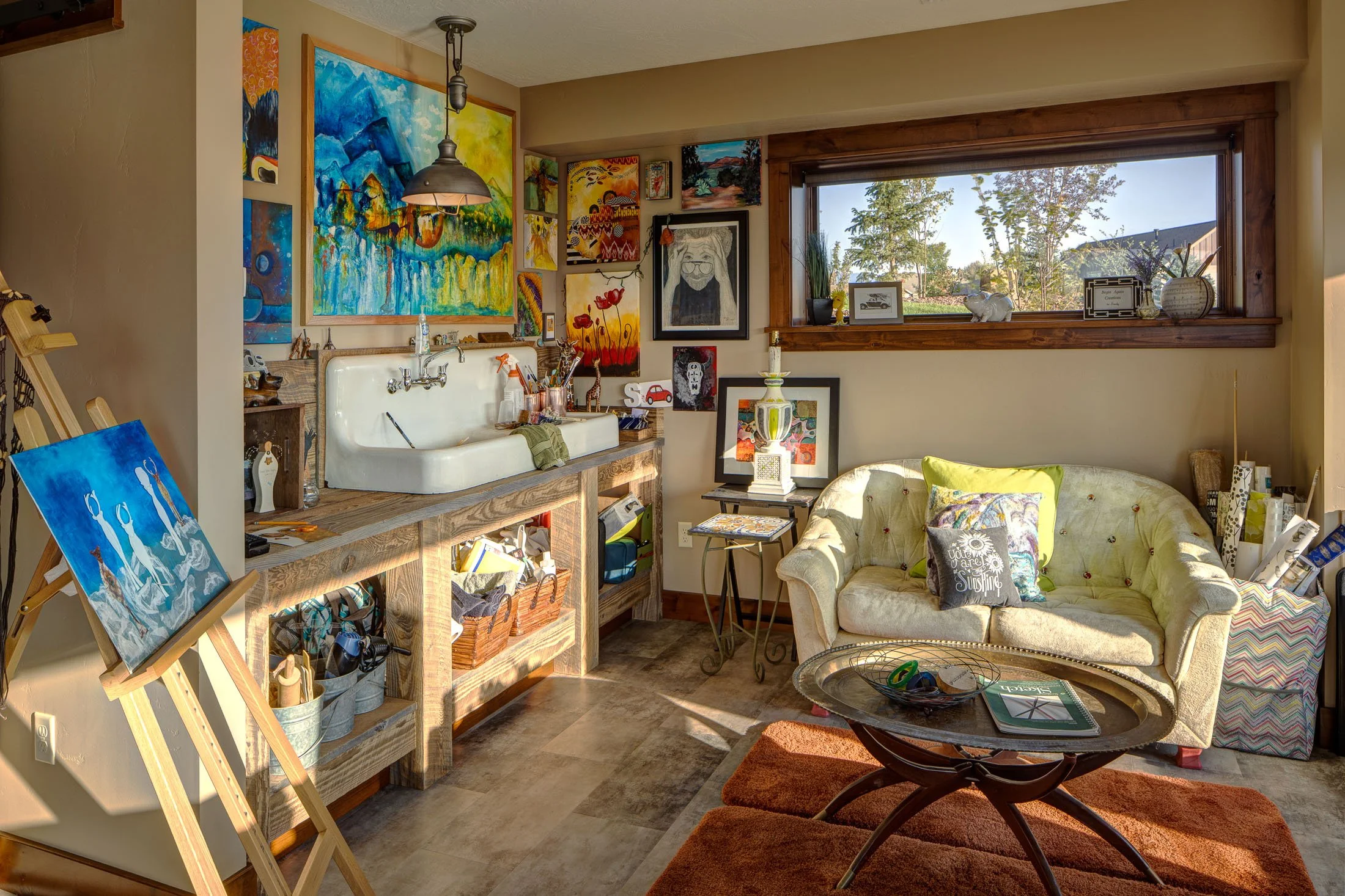 Living room with artwork on walls, a white sofa with pillows, a wooden coffee table, a window viewing trees outside, and an art easel with a painting. Custom Home built in Montana by Edgell Building.