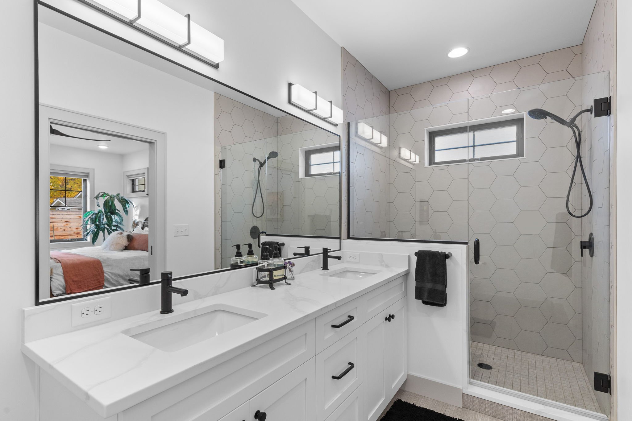 Modern bathroom with double sink vanity, large mirror, black fixtures, and glass shower with hexagonal tiles, small window, and a towel hanging on a rack. Custom Home built in Montana by Edgell Building.