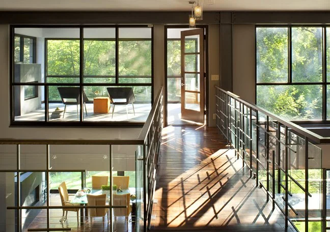 Sunlit modern apartment interior with large windows, a balcony with chairs and a small table, and a staircase leading to a lower level with a dining area. Custom Home built in Montana by Edgell Building.