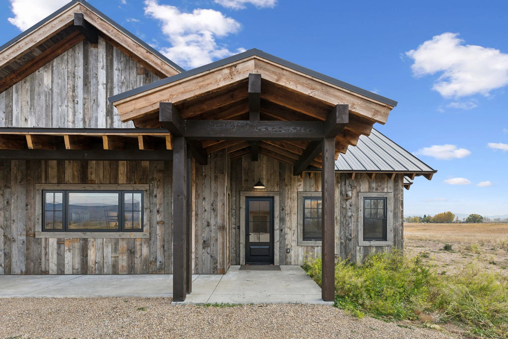 A rustic wooden house with vertical weathered wood siding, black-framed windows, and a front porch covered by a gabled roof with black brackets. The house is set in a rural landscape with fields. Custom Home built in Montana by Edgell Building.