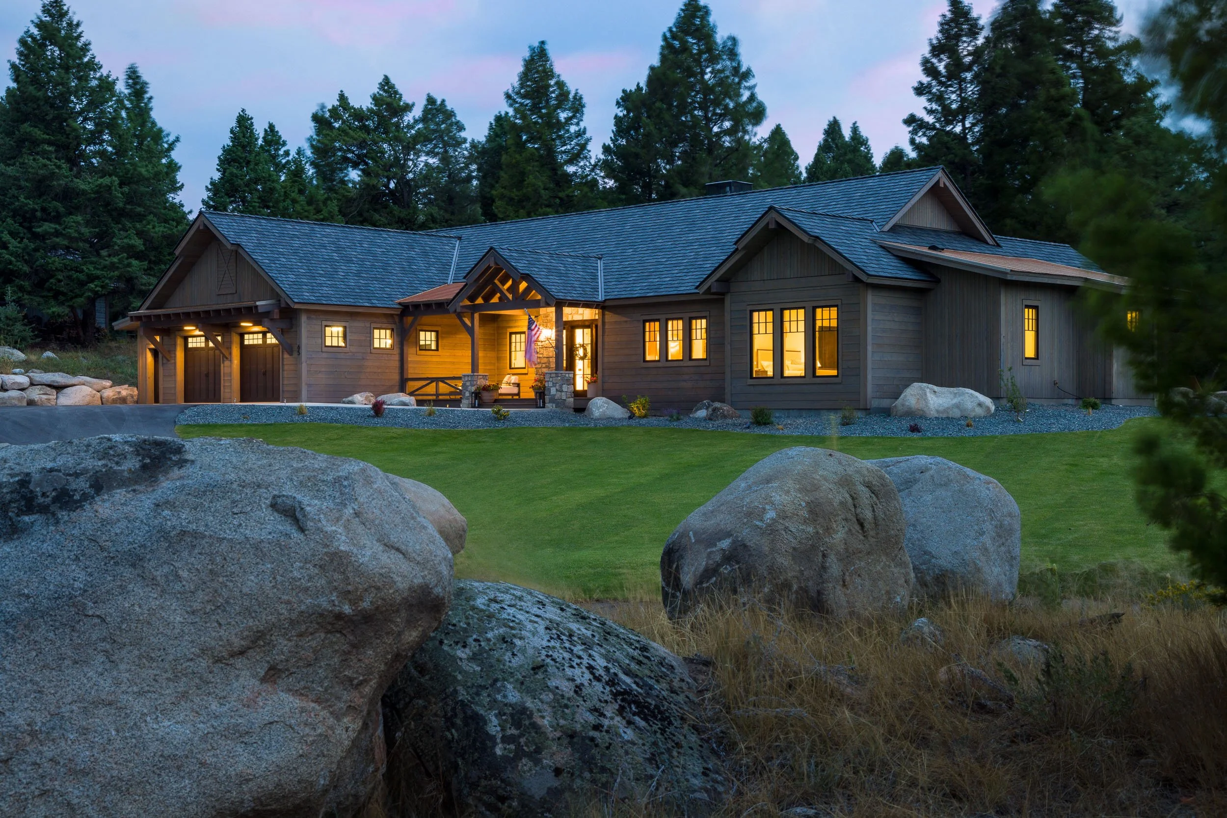 A large, modern house with wooden exterior illuminated from within, surrounded by trees, rocks, and well-manicured lawn at dusk. Custom Home built in Montana by Edgell Building.
