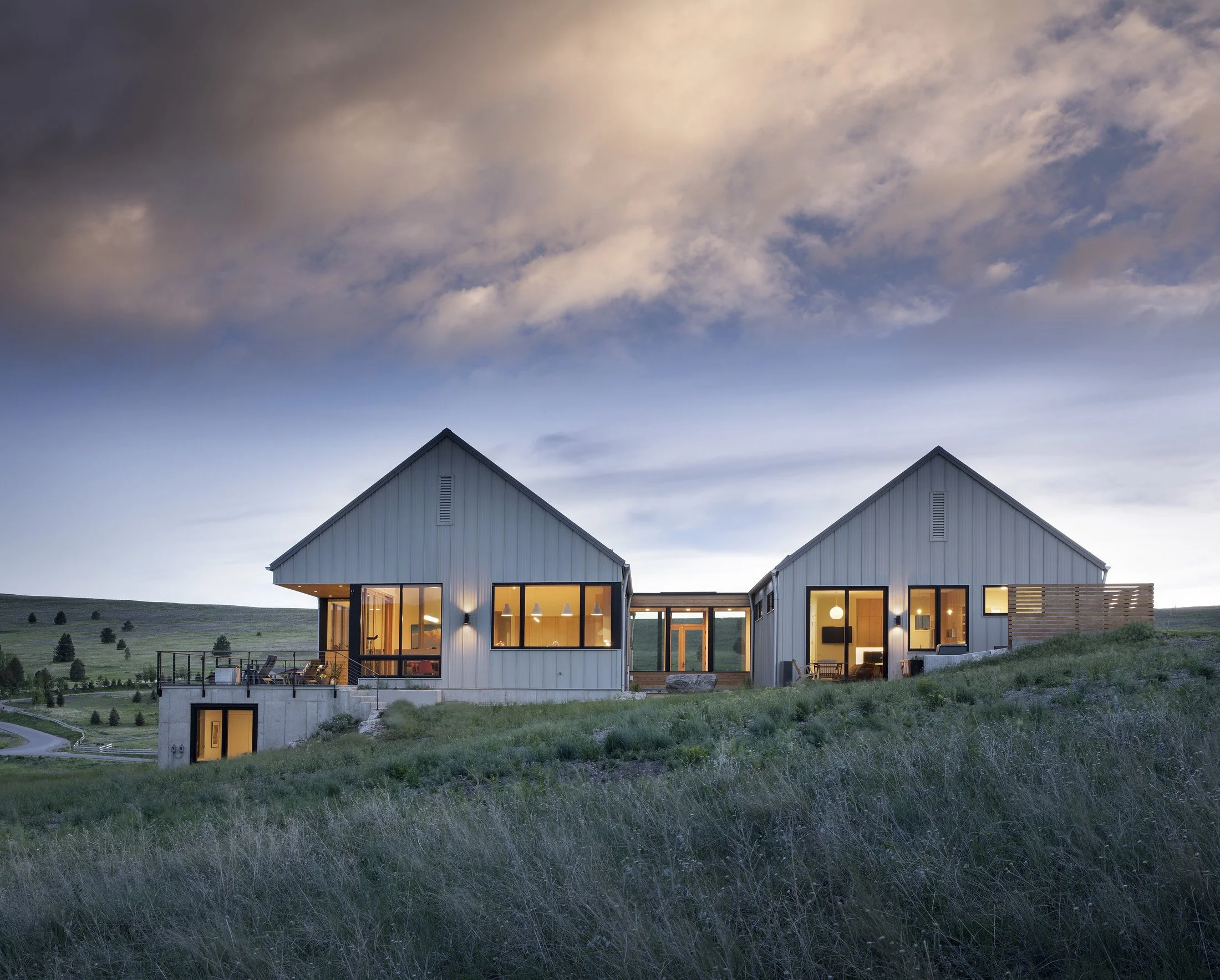 Two modern houses with metal siding and large windows on a grassy hillside at dusk, with a cloudy sky overhead. Custom Home built in Montana by Edgell Building.
