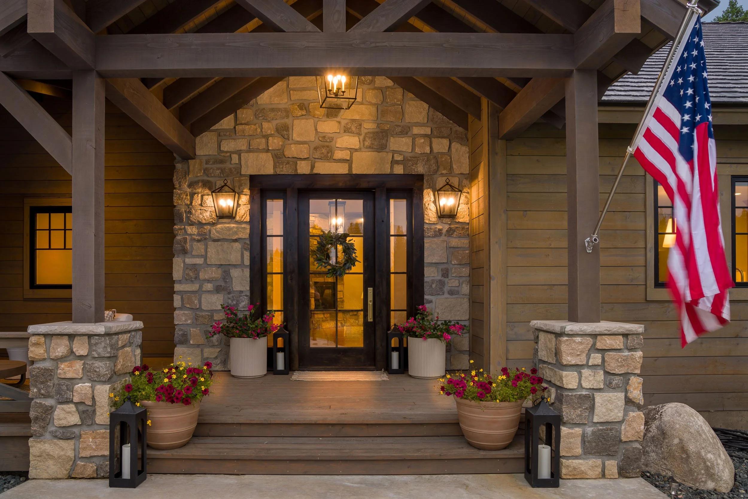 Front porch of a house with stone and wood exterior, decorated with potted flowers, lanterns, and a wreath on the door, and an American flag hanging on the right side. Custom Home built in Montana by Edgell Building.