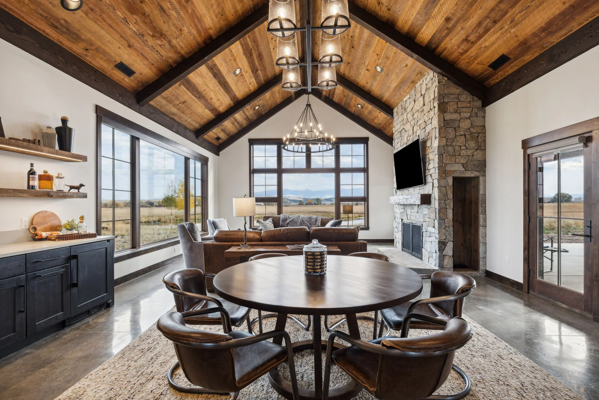 Living room with vaulted wooden ceiling, large windows overlooking a field, stone fireplace with mounted TV, round dining table with six chairs, leather and fabric sofa, side tables, and shelves.  Custom Home built in Montana by Edgell Building.