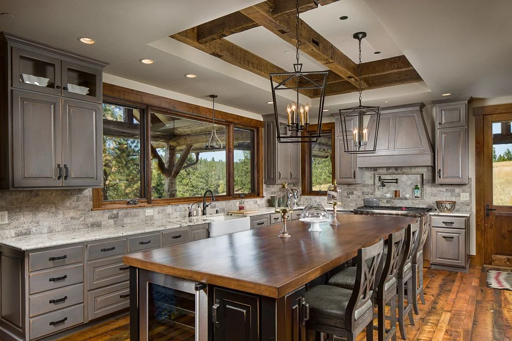Kitchen with gray cabinets, wooden kitchen island, large window, wooden beams, pendant lighting, and a view of trees outside. Custom Home built in Montana by Edgell Building.