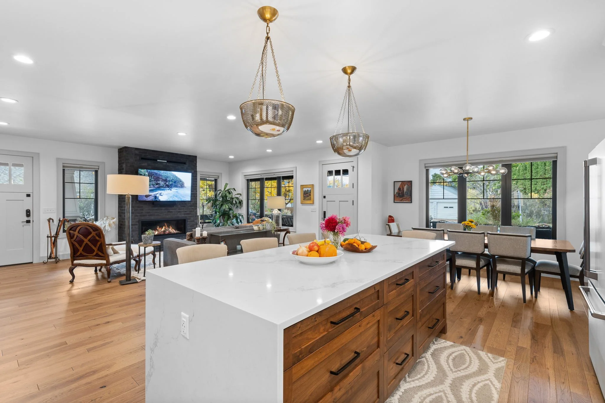 Open-concept living room and kitchen with white walls, hardwood floors, central island, dining table with chairs, fireplace, large windows, and modern lighting fixtures. Custom Home built in Montana by Edgell Building.