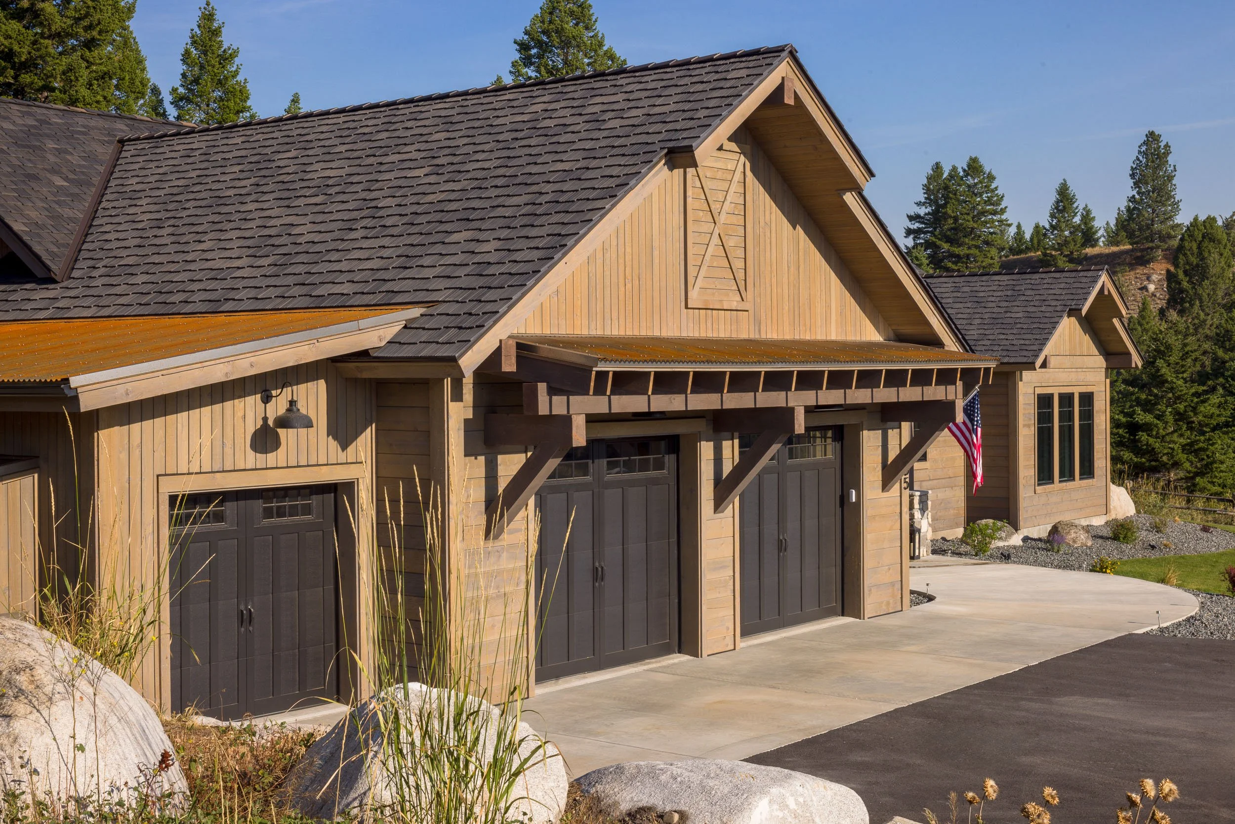 A modern house with a wooden exterior, black garage doors, and a sloped roof, surrounded by greenery and trees in the background. Custom Home built in Montana by Edgell Building.