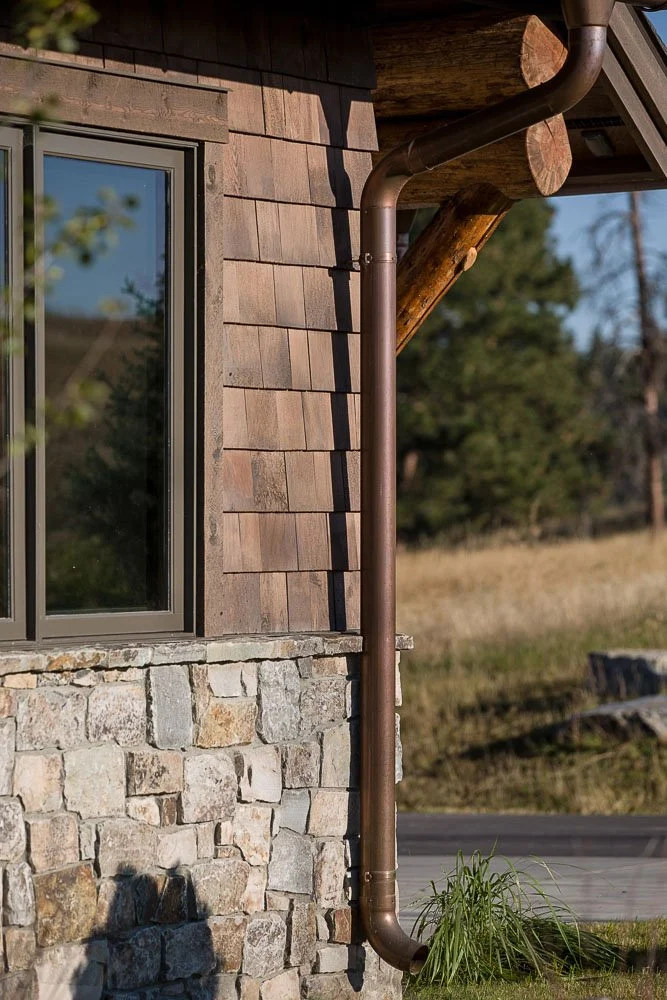 Close-up of a house corner showing a window, wooden shingles, stone foundation, and a metal downspout with a grassy plant at its base, outdoor rural setting. Custom Home built in Montana by Edgell Building.