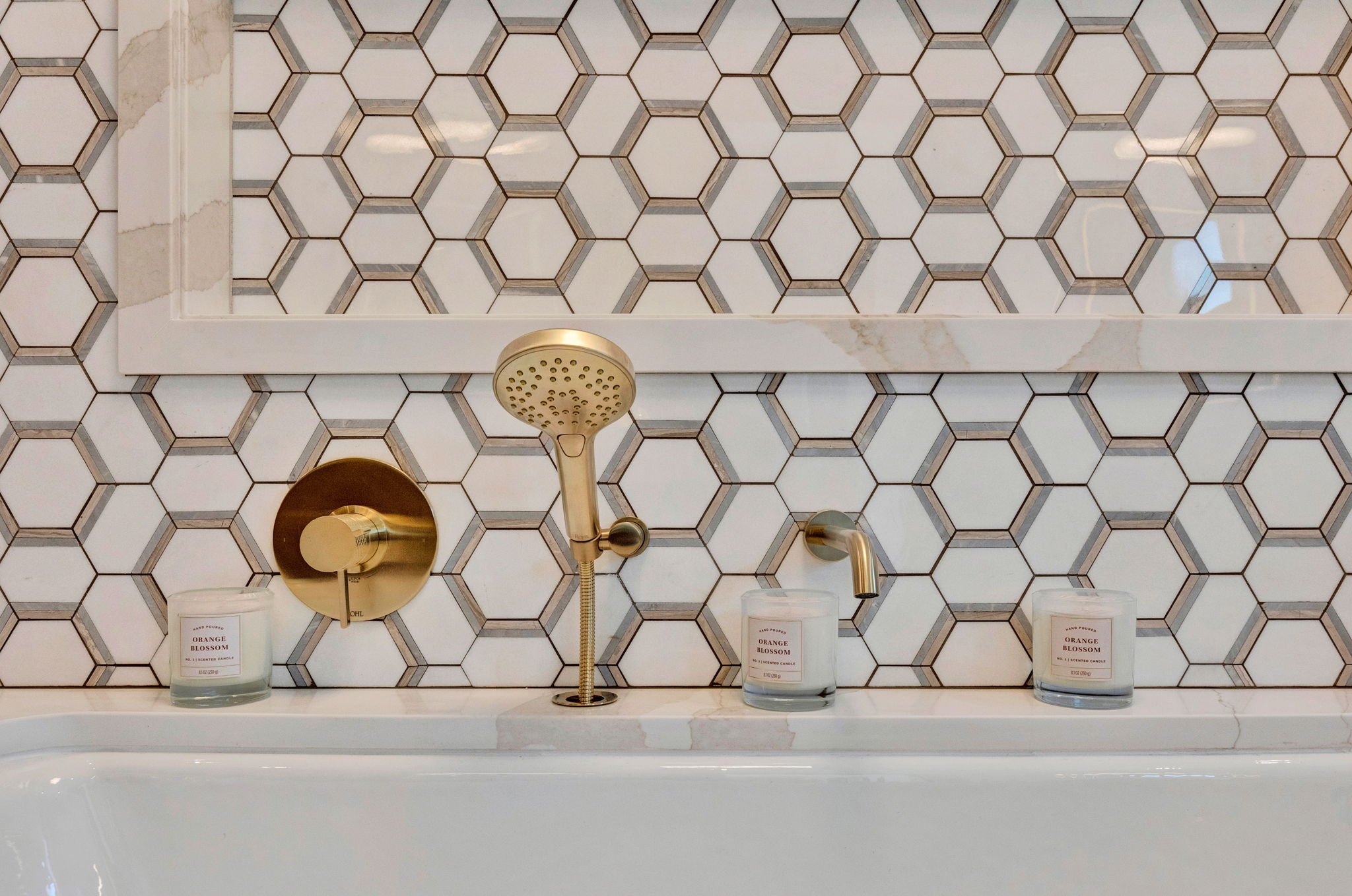 Close-up of a bathroom wall with white hexagonal tiles and brass fixtures, including a handheld showerhead, a wall-mounted faucet, and two candles labeled 'Orange Blossom' on a white bathtub edge. Custom Home built in Montana by Edgell Building.