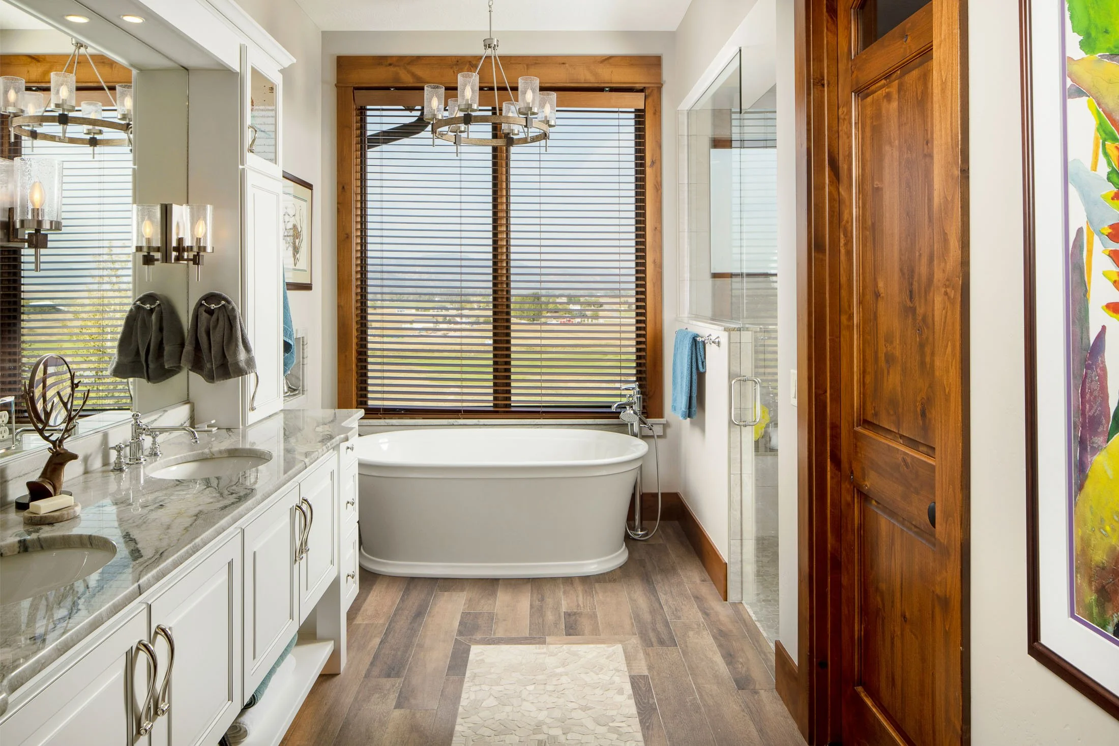 Bathroom with double vanity sinks, marble countertops, a large mirror, a freestanding bathtub, wooden window blinds, a shower stall, wooden cabinets, and colorful artwork on the wall. Custom Home built in Montana by Edgell Building.