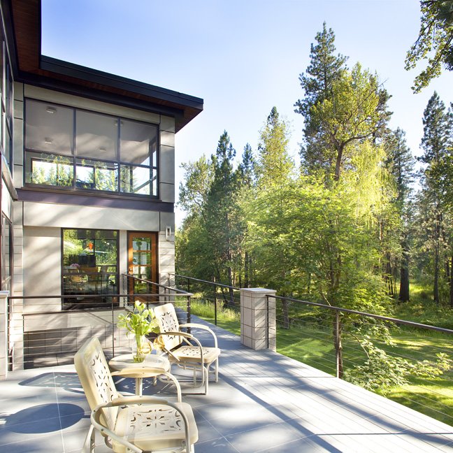 Modern house with large glass windows, a spacious balcony with chairs and potted plants, surrounded by green trees and a clear sky. Custom Home built in Montana by Edgell Building.