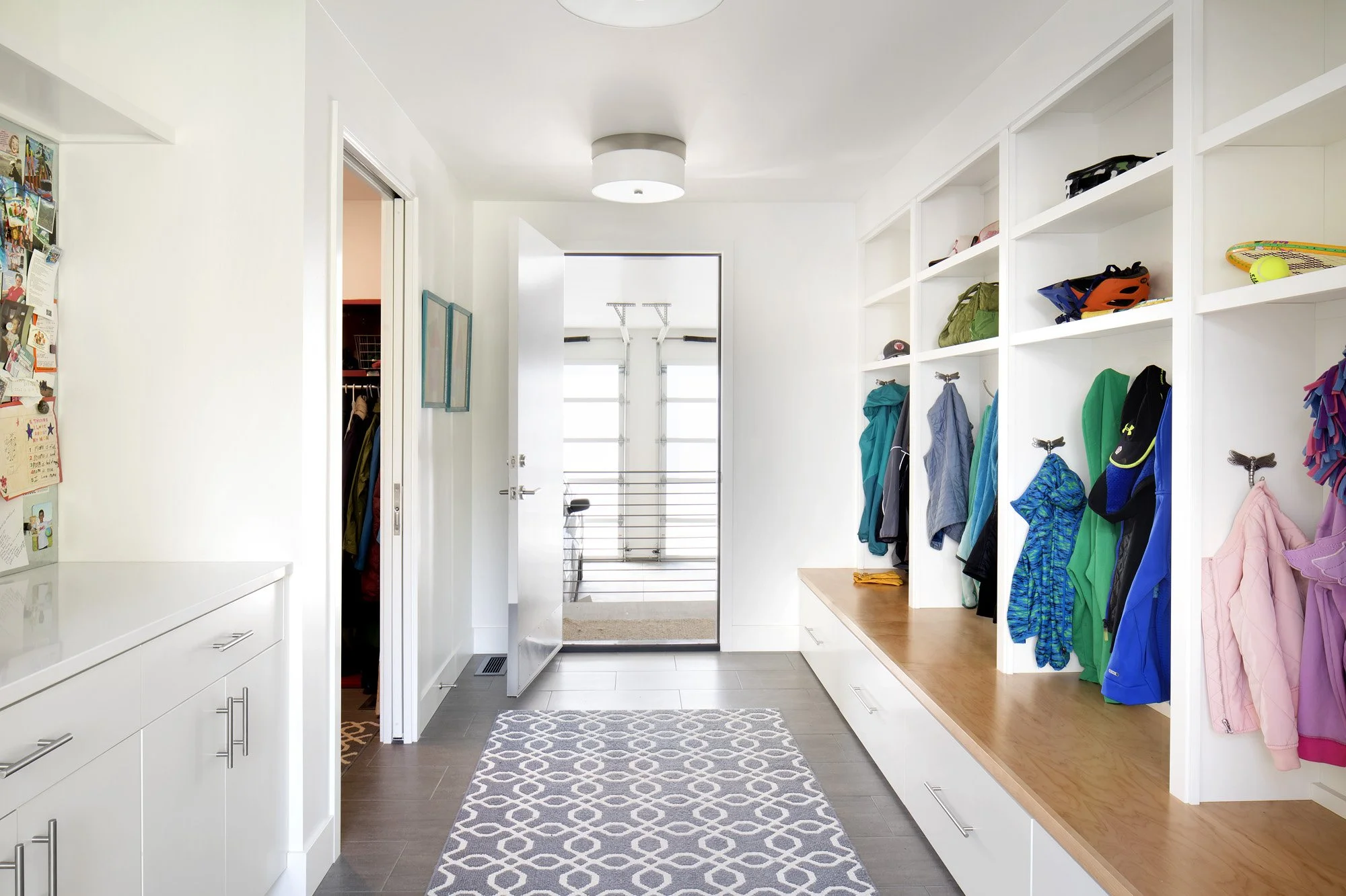 Bright and organized front entryway with white built-in cubbies holding jackets, hats, and helmets, a bench with storage drawers, a patterned rug, and a view through the open door to a staircase. Custom Home built in Montana by Edgell Building.