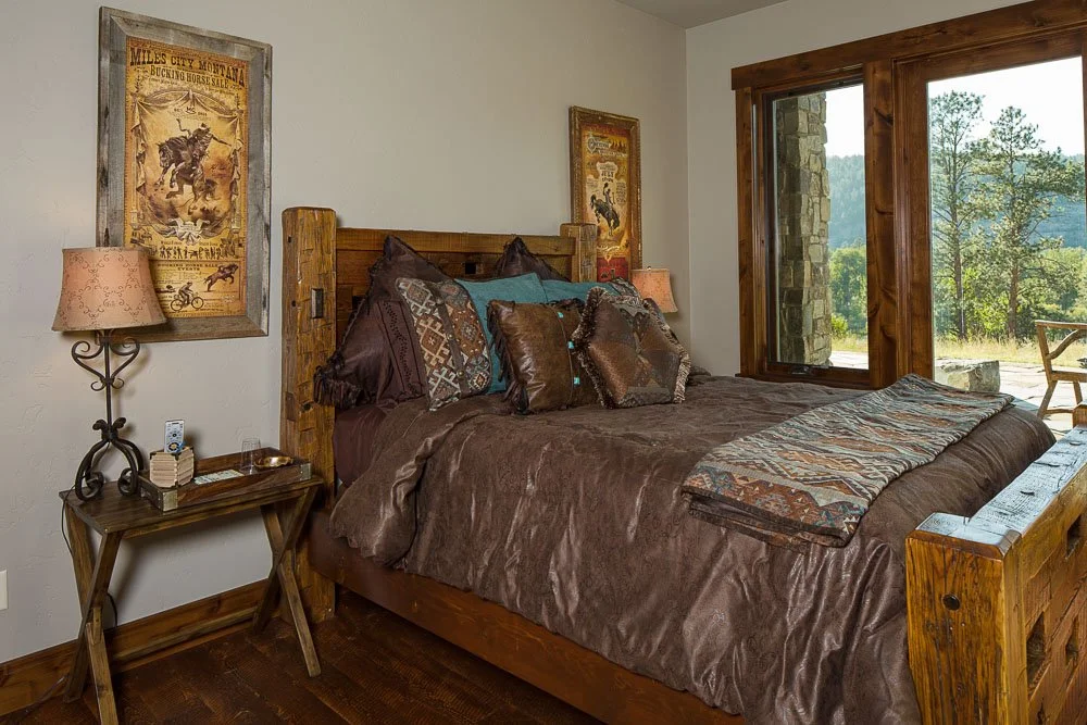 Cozy bedroom with wooden furniture, brown bedding, decorative pillows, and wall art featuring vintage western posters. Large window with a mountain view, next to a small wooden table holding a lamp and remote control. Custom Home built in Montana by 