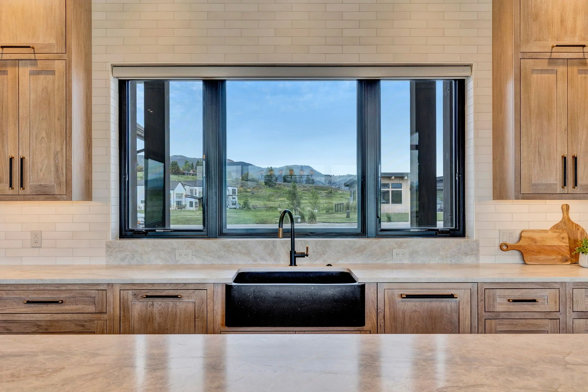 Modern kitchen with black sink, wooden cabinets, and large window showing a scenic view of trees, houses, and mountains. Custom Home built in Montana by Edgell Building.