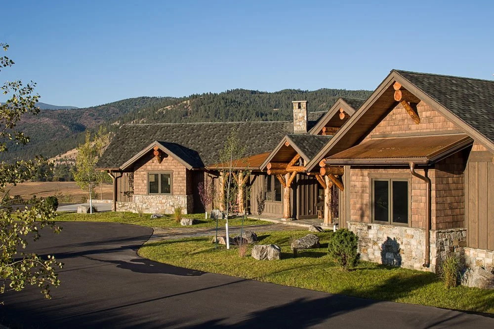 A row of rustic mountain houses with wood and stone exteriors, surrounded by green lawns and young trees, against a backdrop of forested hills and mountains under a clear blue sky. Custom Home built in Montana by Edgell Building.