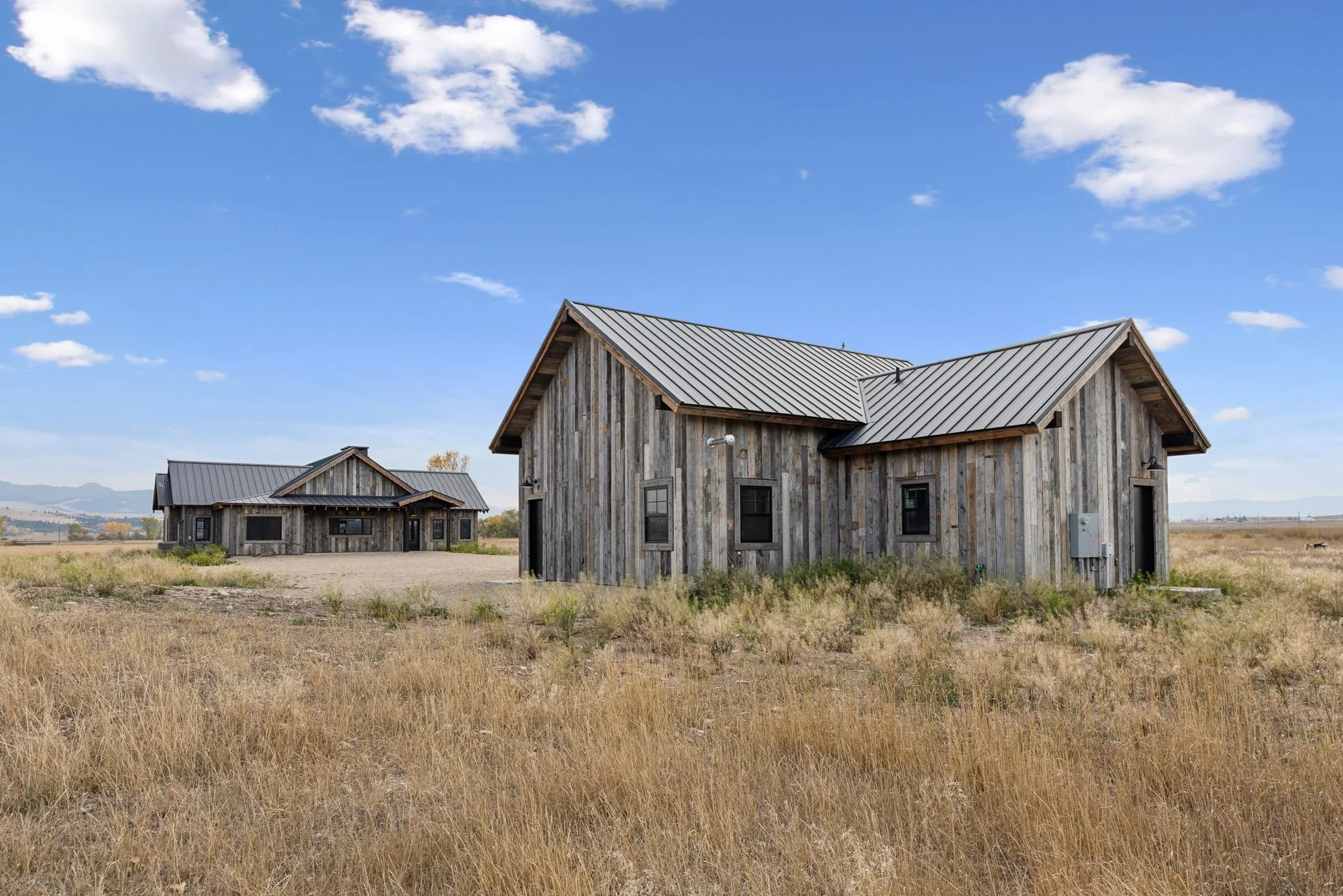 Two rustic wooden houses in a flat grassy field under a blue sky with scattered clouds. Custom Home built in Montana by Edgell Building.