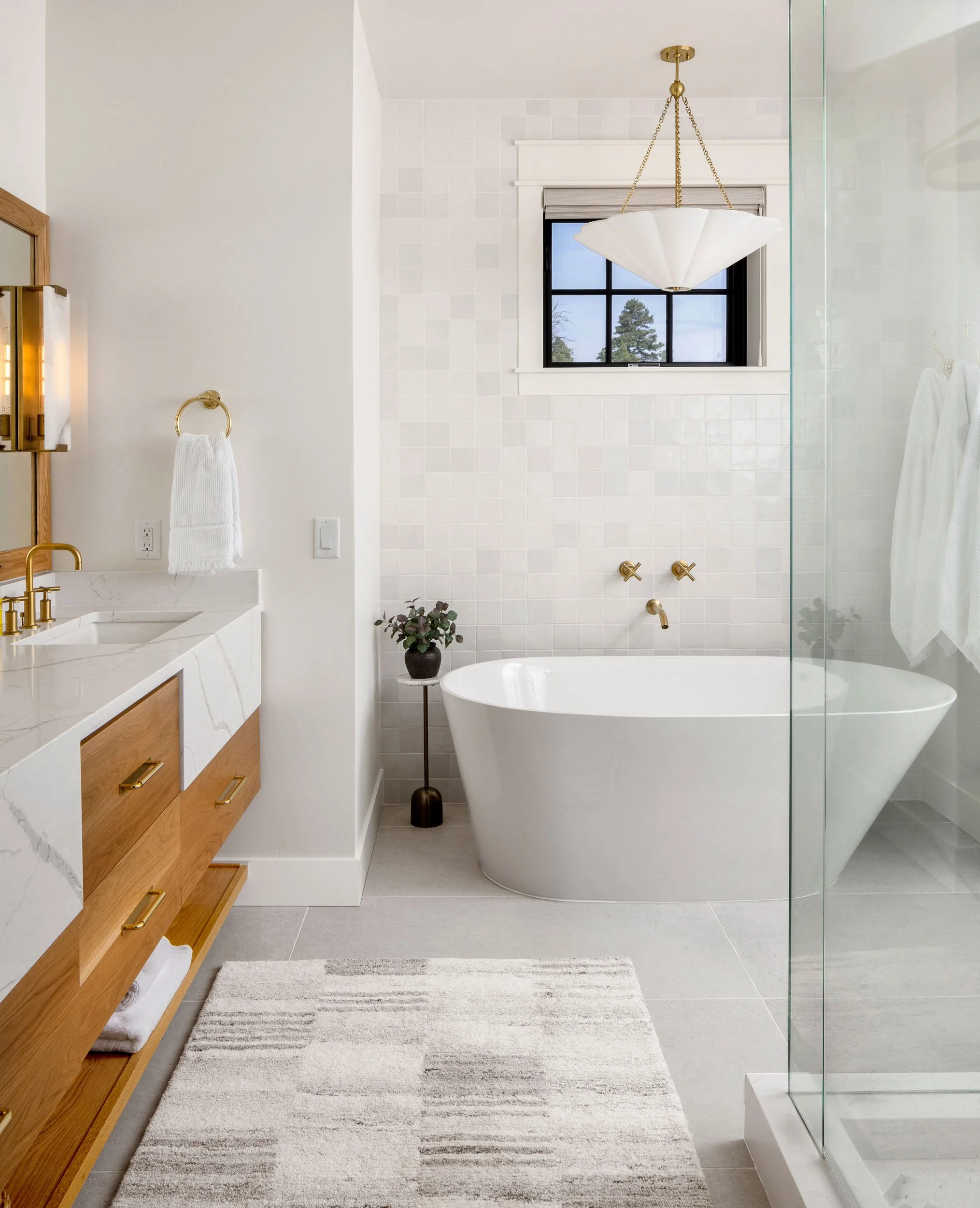 Modern bathroom with white freestanding bathtub, small window, white tiled wall, gold fixtures, wooden vanity with white marble countertop, towel rack, potted plant, and a striped rug. Custom Home built in Montana by Edgell Building.