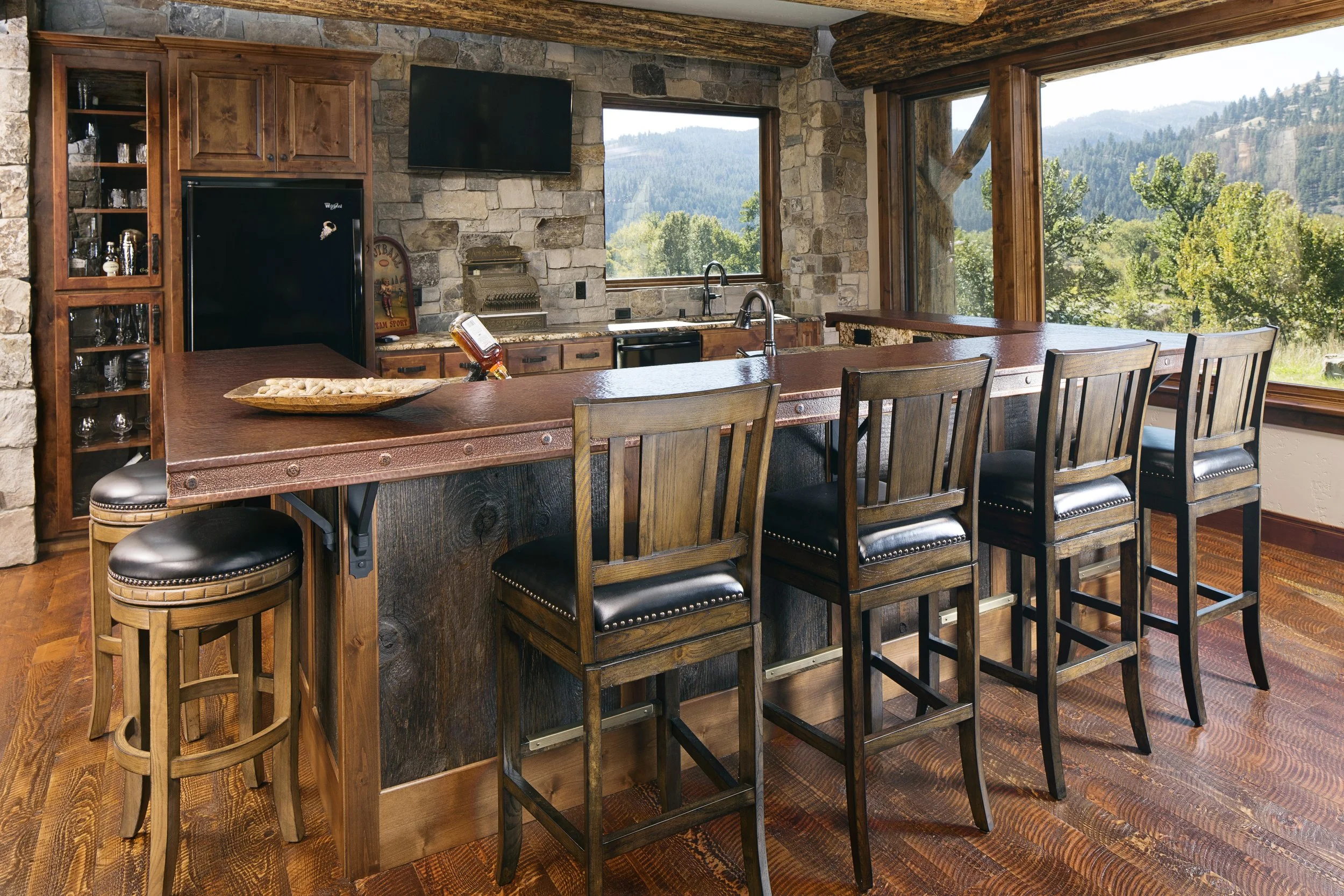 A rustic kitchen with a large wooden island, four black leather cushioned bar stools, stone walls, large windows showing mountain views, and wooden beams on the ceiling. Custom Home built in Montana by Edgell Building.