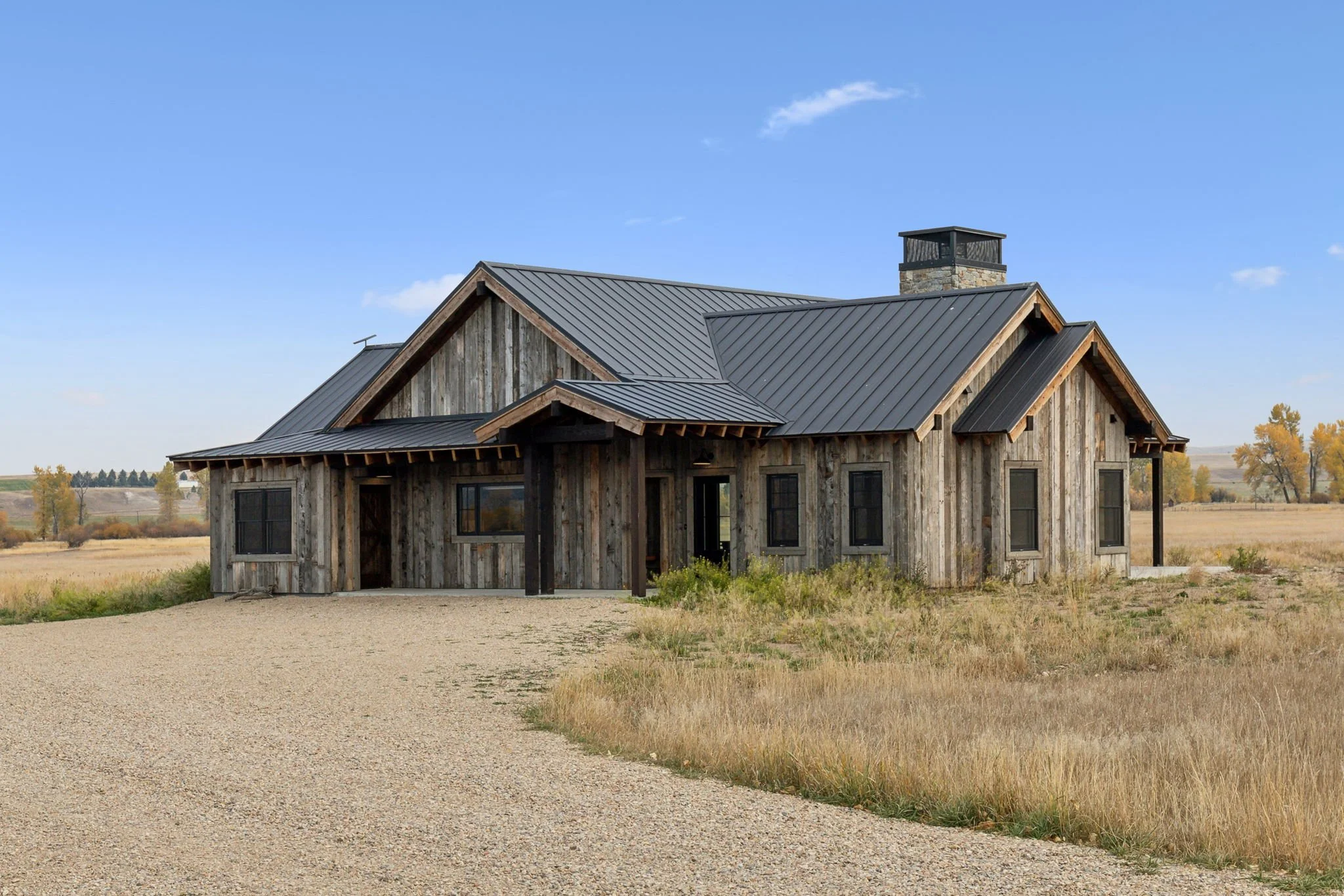 A rustic house with wooden siding and a metal roof, situated in a rural area with a gravel driveway and dry grasslands, under a blue sky.