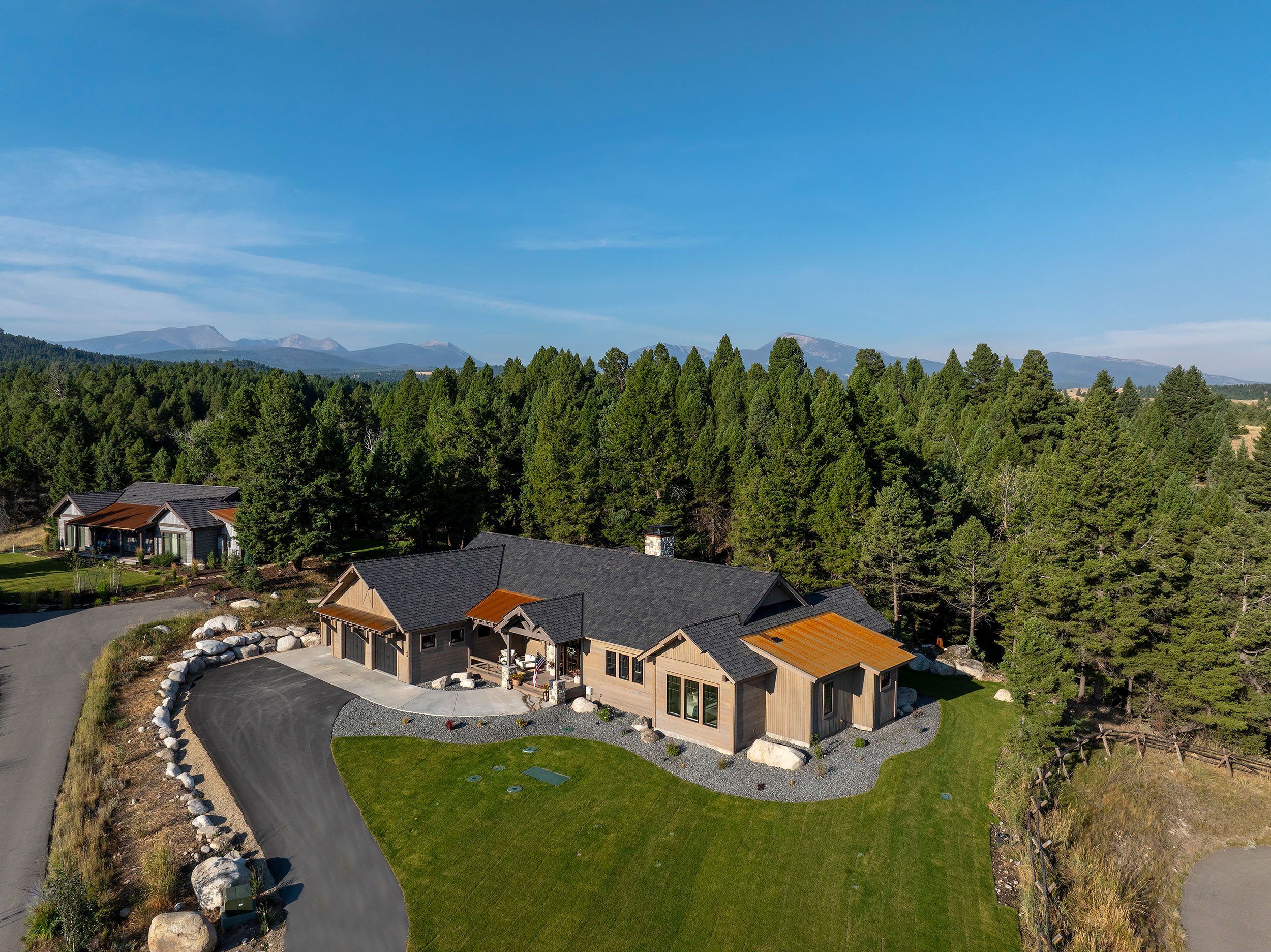 A house with a black roof and wooden siding surrounded by trees and a green lawn, with a gravel driveway in front, set in a mountainous landscape under a blue sky. Home Designed and Built By Edgell Building in Western Montana.