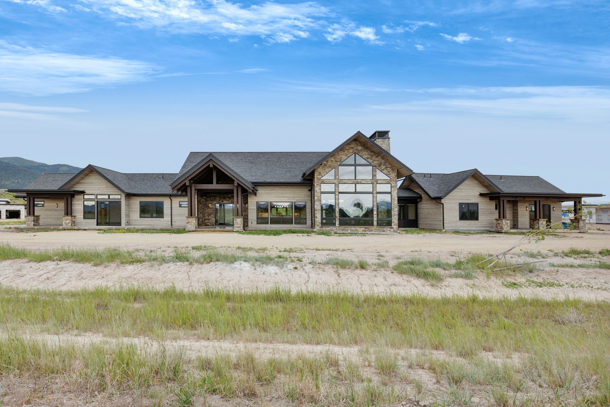 A large modern house under construction with a mostly finished exterior, large windows, and a combination of stone and wood siding. The house is situated in a sandy, grassy area under a blue sky. Custom Home built in Montana by Edgell Building.