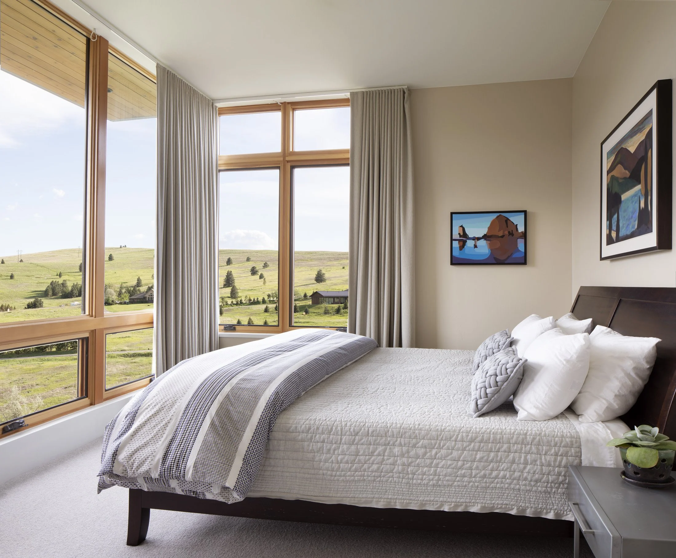 Modern bedroom with a bed near large corner windows showing a green landscape, framed artwork on the walls, and a small nightstand with a potted plant. Custom Home built in Montana by Edgell Building.