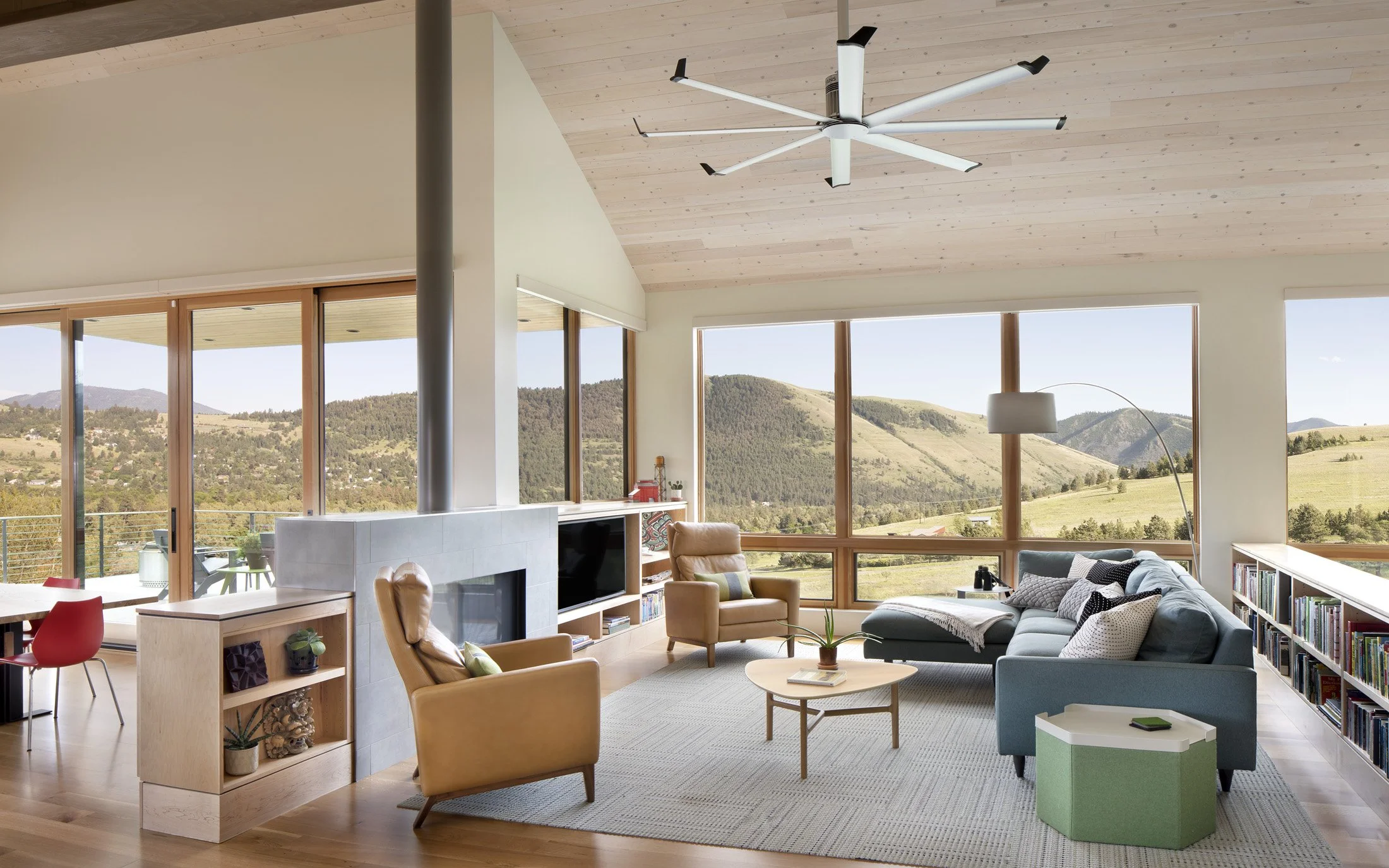 Bright living room with large windows showing scenic mountain landscape, modern furniture including a gray sectional sofa, leather armchair, round coffee table, floor lamp, and built-in bookshelf. Custom Home built in Montana by Edgell Building.