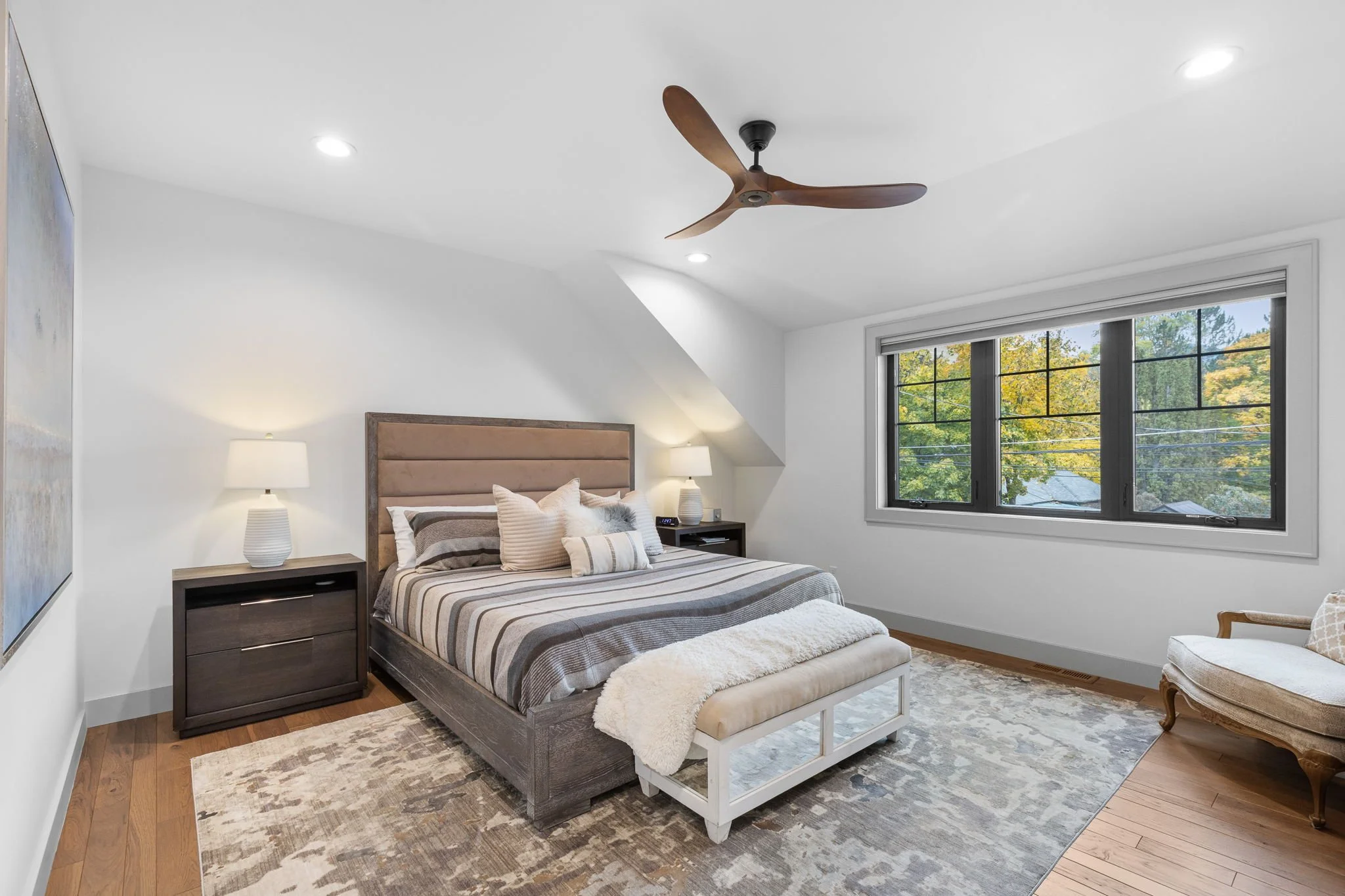 Modern bedroom with a large bed, beige headboard, striped bedding, white throw, nightstands with lamps, a window with trees outside, a ceiling fan, and an armchair. Custom Home built in Montana by Edgell Building.