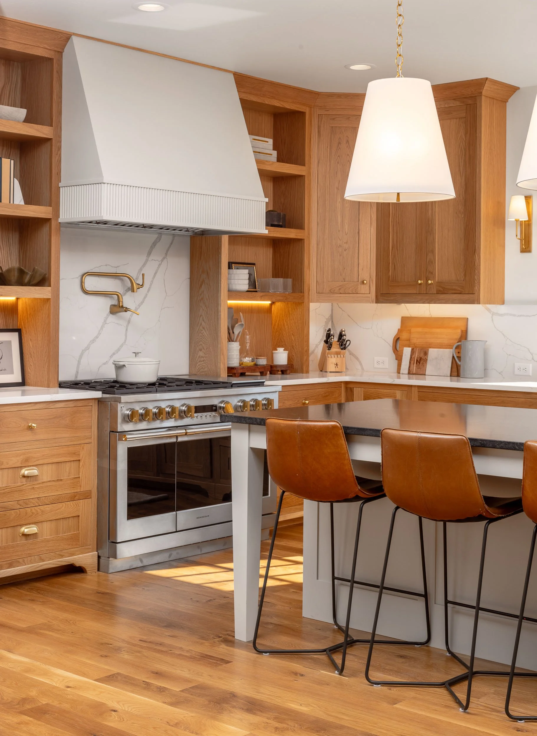 A modern kitchen with wooden cabinetry, a white marble backsplash, a stainless steel oven, a white range hood, brown leather bar stools, and pendant lighting. Custom Home built in Montana by Edgell Building.