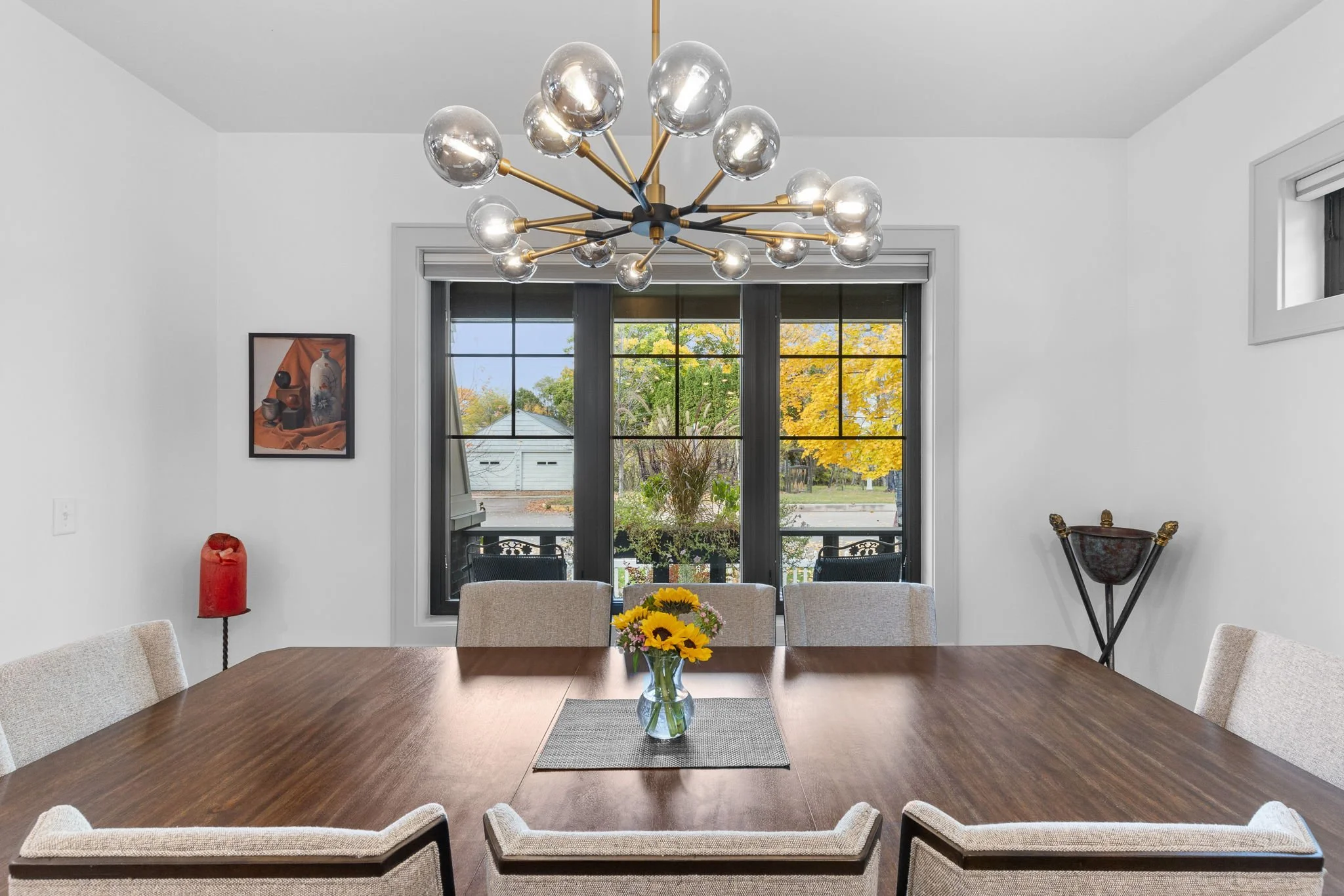 Modern dining room with a large wooden table and beige chairs, a contemporary chandelier overhead, a window with a view of trees with fall foliage, and decorative items including a flower vase. Custom Home built in Montana by Edgell Building.