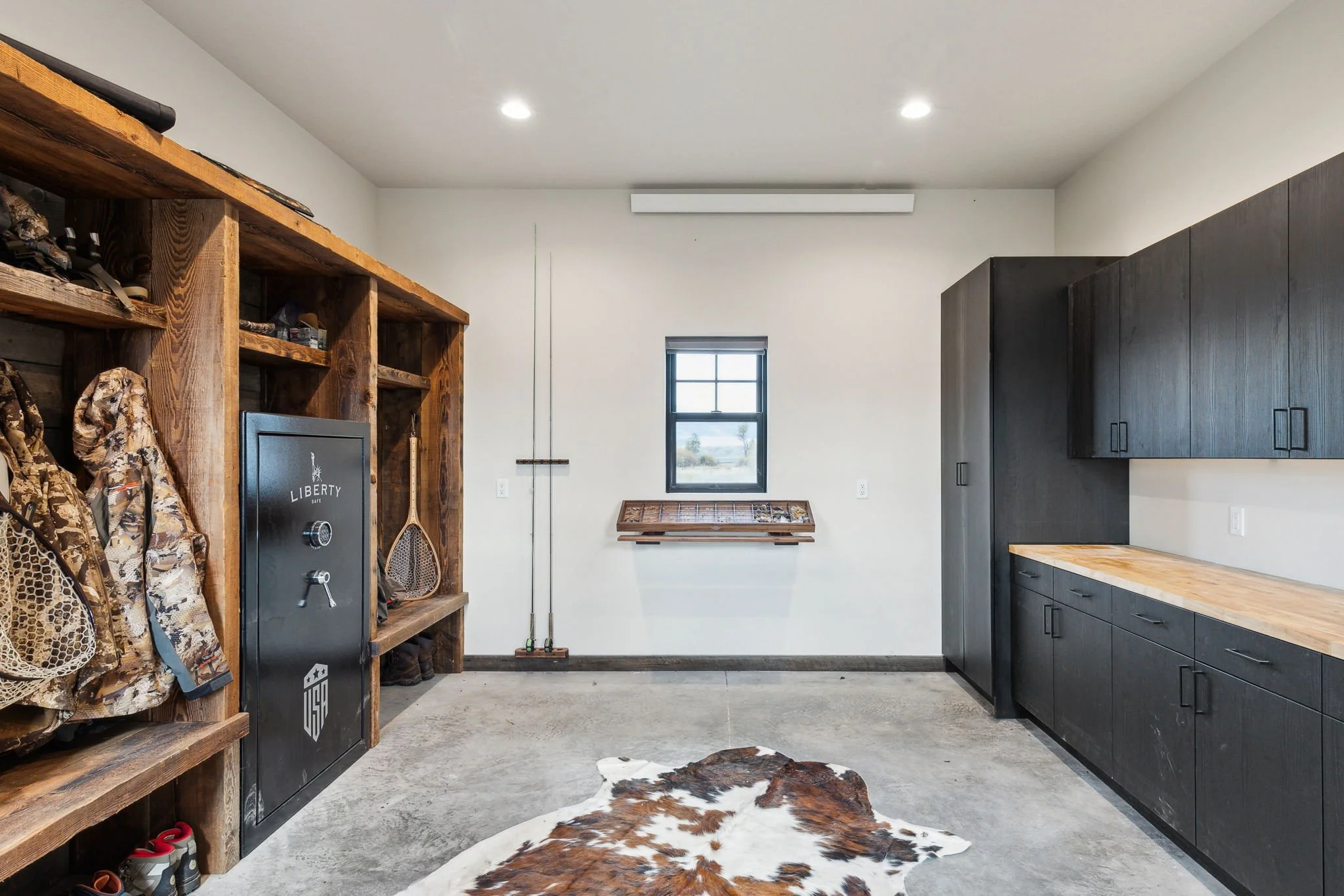 Garage with black storage cabinets, wooden work surface, wooden shelves, a safe, and hunting gear including camouflage clothing and rifles, with a cowhide rug on the concrete floor and a small window. Custom Home built in Montana by Edgell Building.