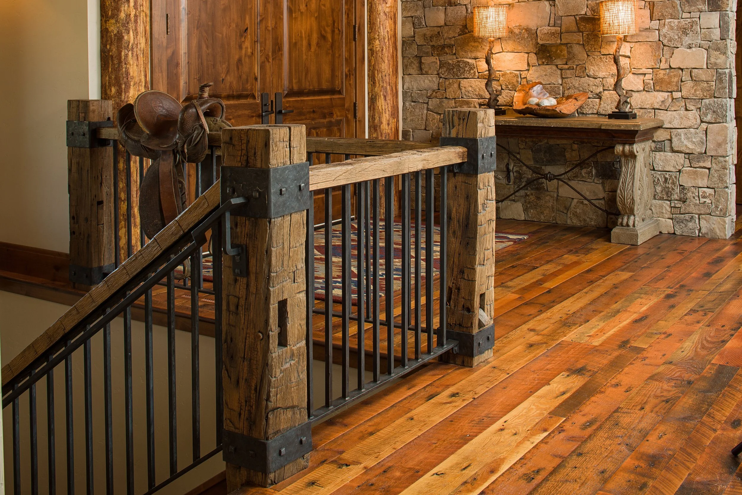 Rustic interior with a wooden railing, a stone wall, and a wood console table with decorative objects and lamps. Custom Home built in Montana by Edgell Building.