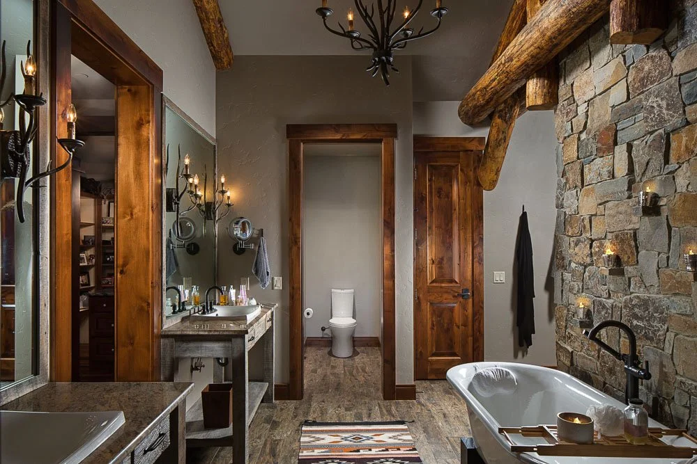 Rustic bathroom with stone accent wall, wooden beams, and a clawfoot tub. There is a toilet in a separate small room, a wooden door, a double vanity with mirrors and sconces, and a chandelier. Custom Home built in Montana by Edgell Building.