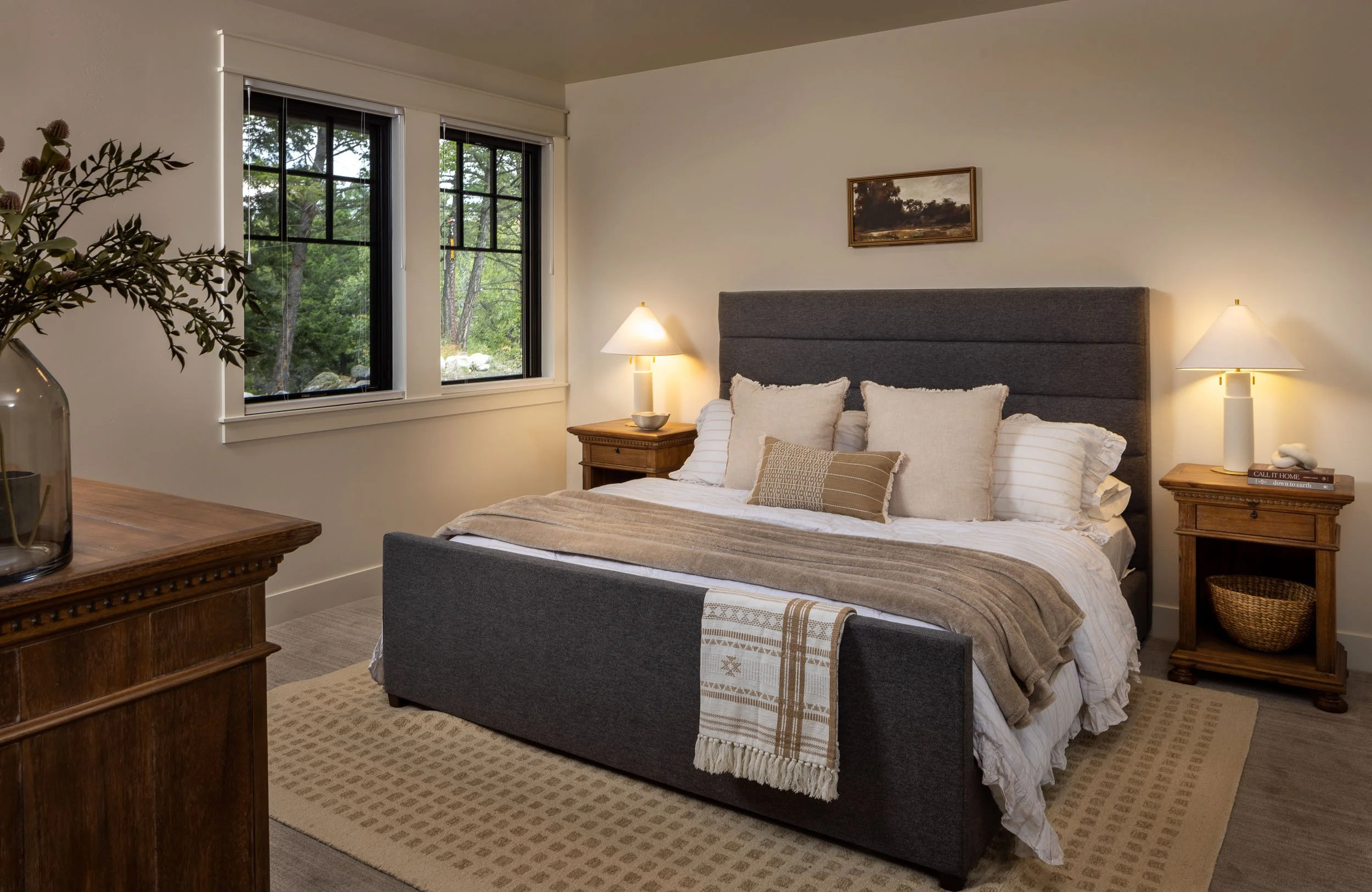 A cozy bedroom with a slate gray bed, beige and white pillows, a beige blanket, and a small decorative pillow. Two wooden nightstands with lamps flank the bed, and a framed landscape painting hangs above. A large window with black frames shows greene