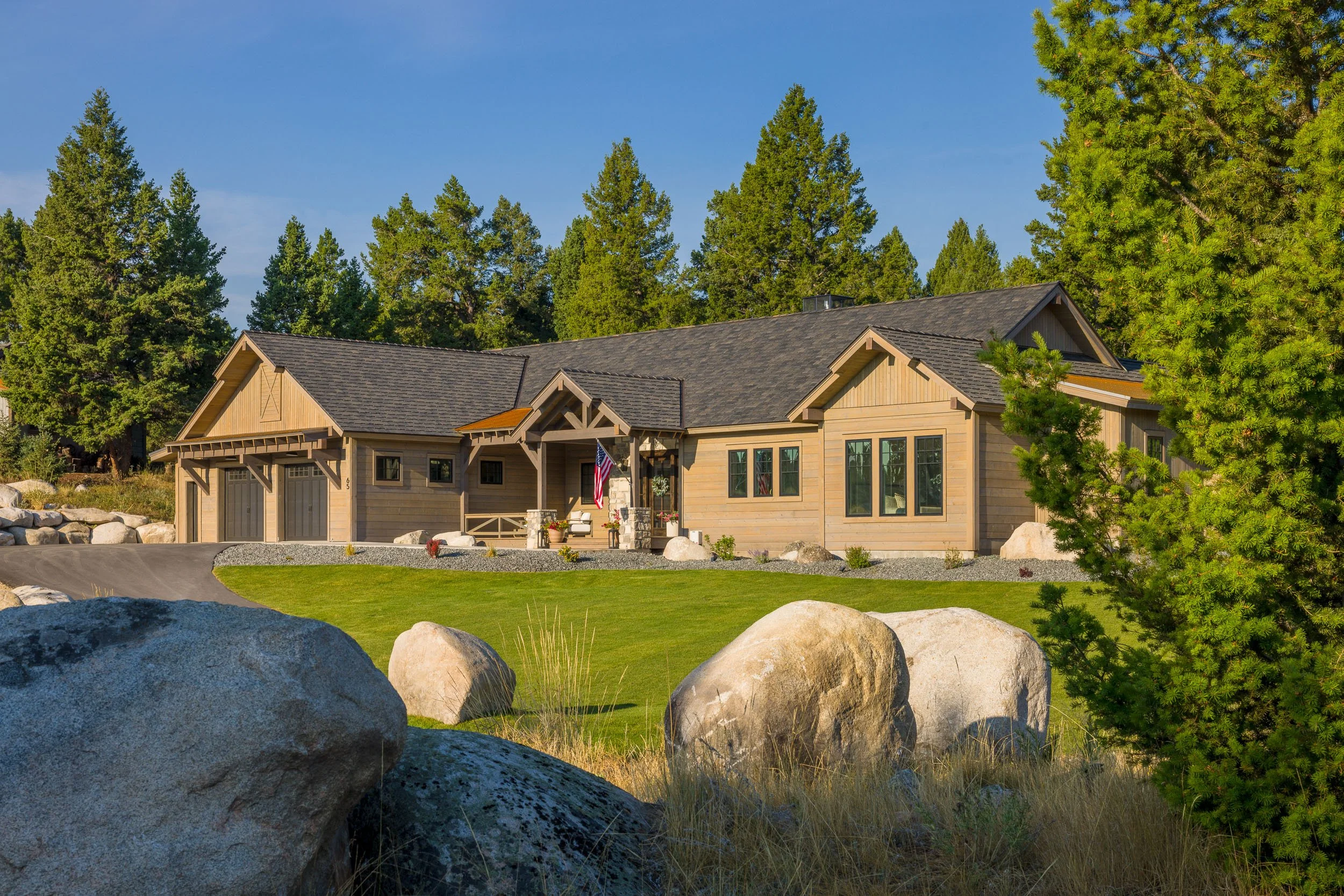 A single-story house with beige wooden siding, dark grey roof, and a small front porch, surrounded by a well-maintained lawn, large rocks, and tall green trees in the background under a clear blue sky. Custom Home built in Montana by Edgell Building.