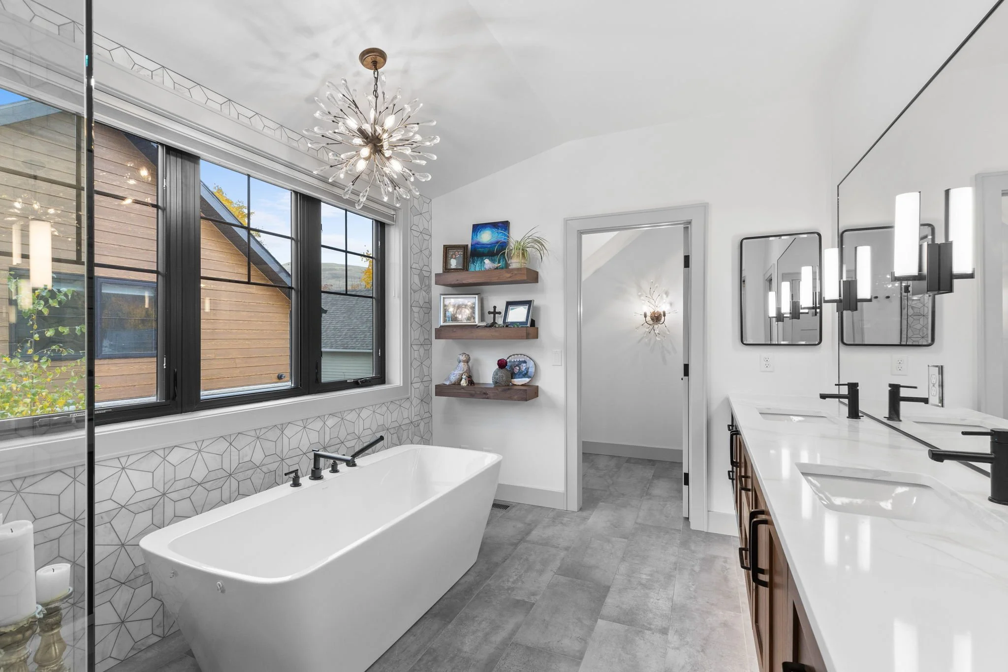 Modern bathroom with white bathtub, large window, gray floor tiles, floating wooden shelves with decor, double vanity with black fixtures, and a unique chandelier. Custom Home built in Montana by Edgell Building.