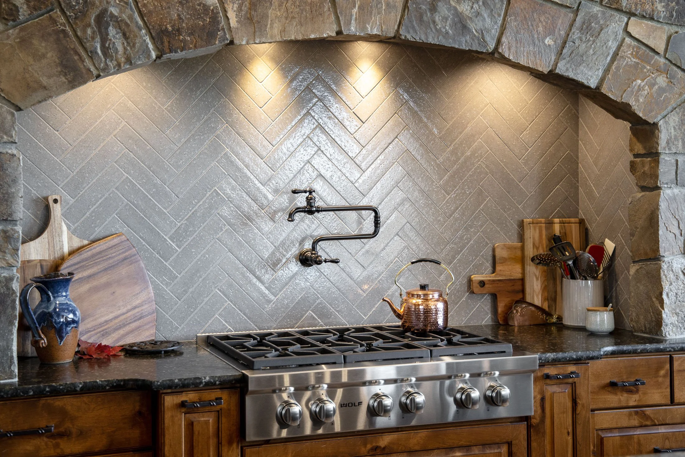 Kitchen with a stainless steel stove, copper teapot, wooden cutting boards, ceramic containers, and utensils, with a stone backsplash and archway. Custom Home built in Montana by Edgell Building.