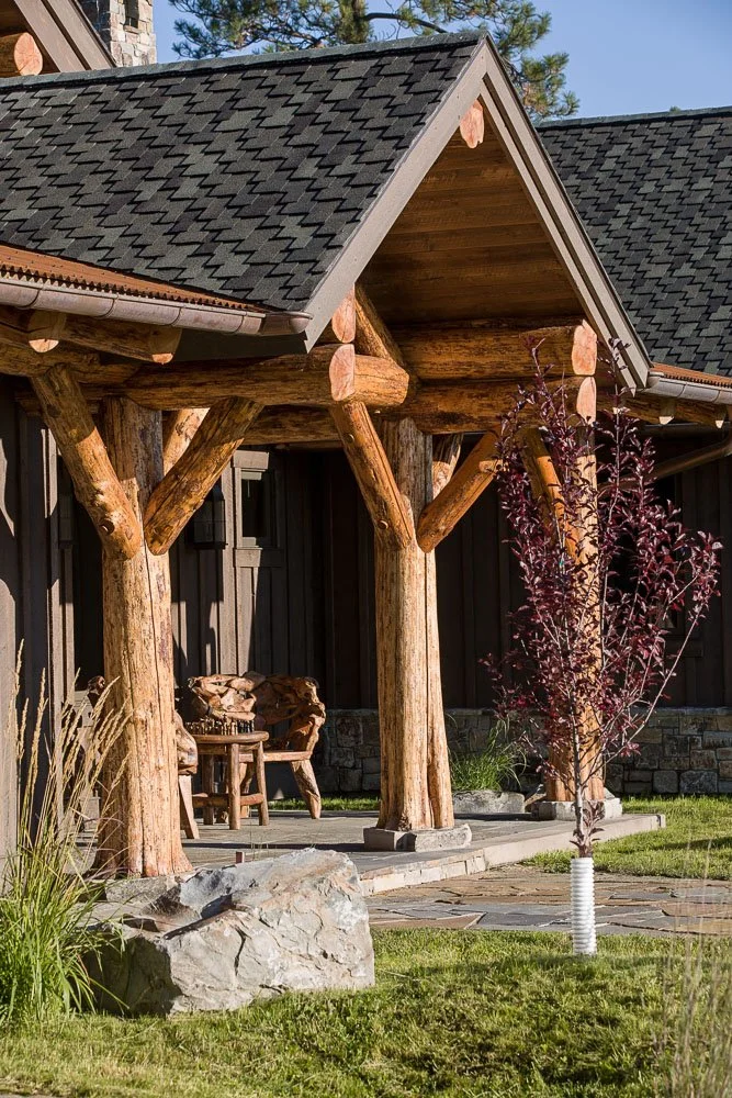 Wooden porch with log supports and a roof, outdoor bench, landscaped yard with rocks and a small tree. Custom Home built in Montana by Edgell Building.