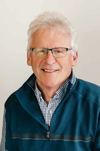 David Edgell of Edgell Building. Portrait of a smiling older man with white hair and glasses, wearing a blue jacket and checkered shirt, standing against a plain light background.