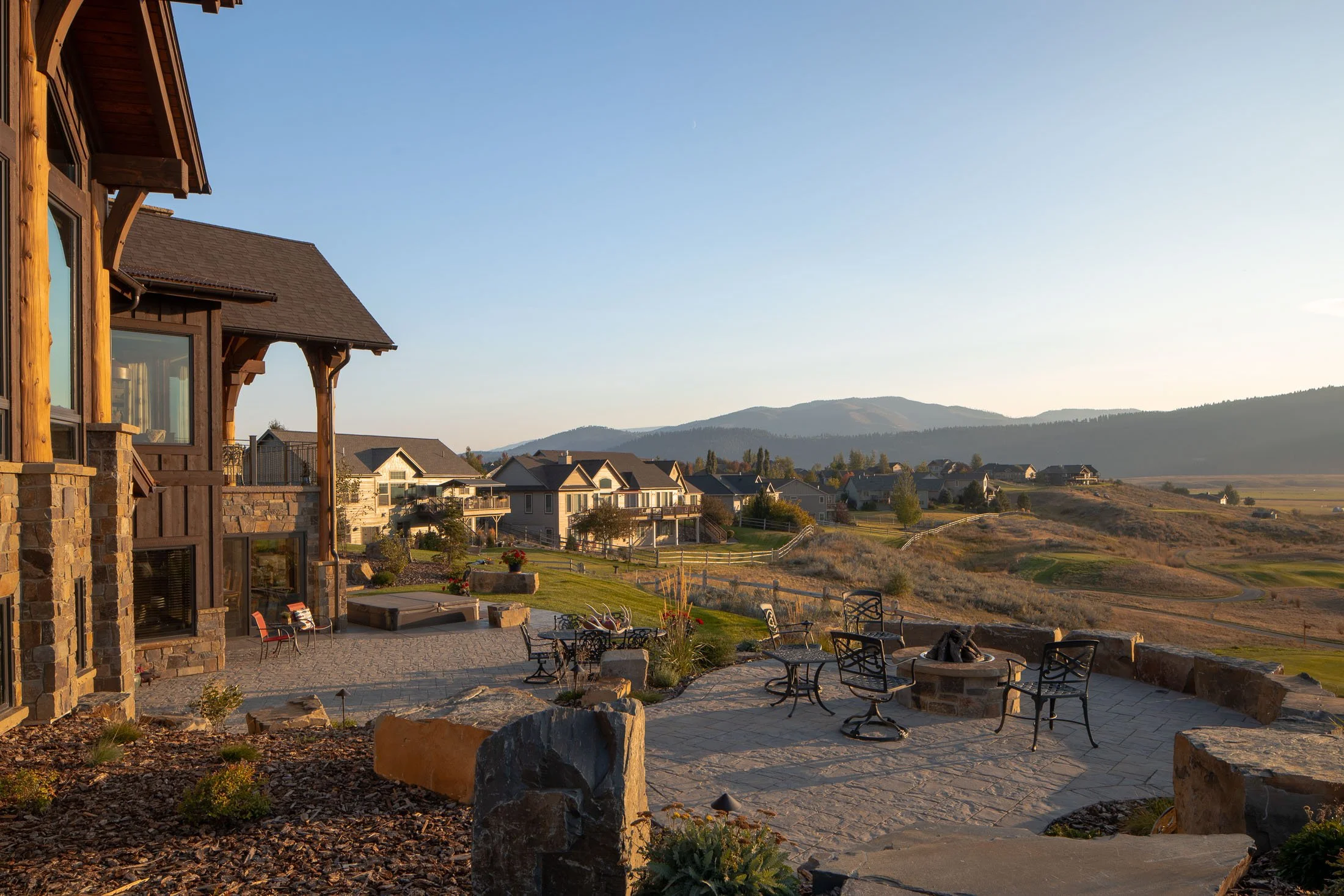 Scenic view from a patio with outdoor seating, overlooking a suburban neighborhood with houses and rolling hills in the background during sunset. Custom Home built in Montana by Edgell Building.